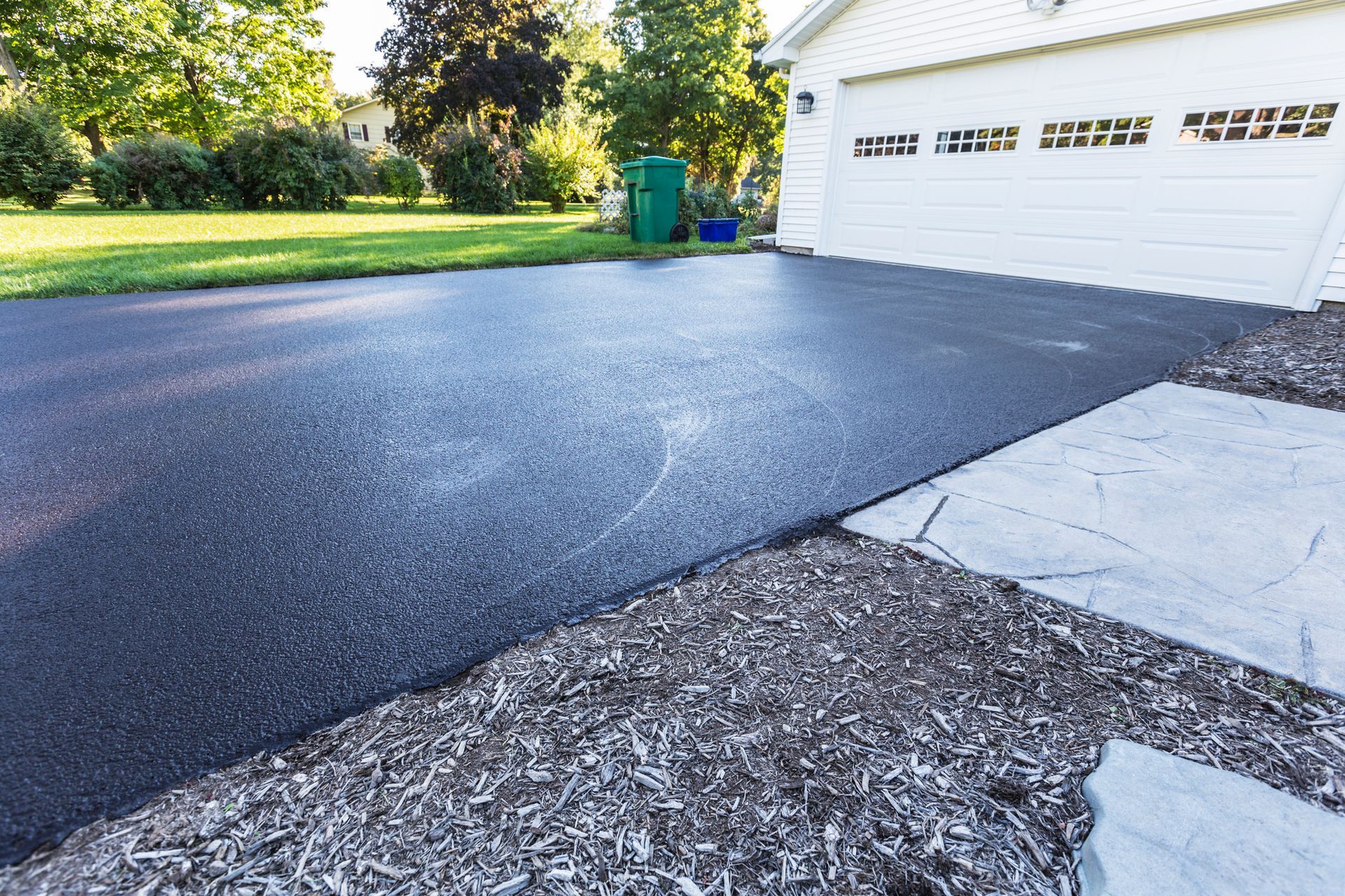 Black asphalt driveway next to a garage, green grass, and a concrete sidewalk.
