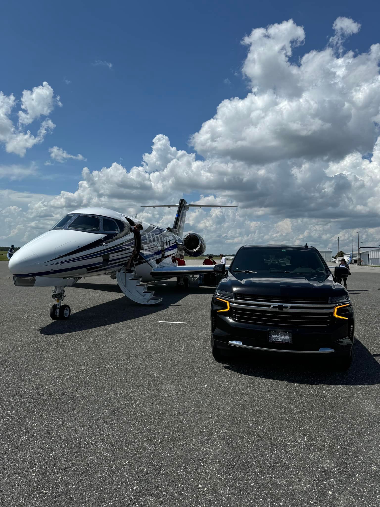 A black SUV parked on an airport tarmac next to a white airplane with a distinctive black patterned livery under blue sky.