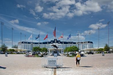 A large, white, multi-story building sits behind a central bronze statue and a row of flags against a blue, cloudy sky.