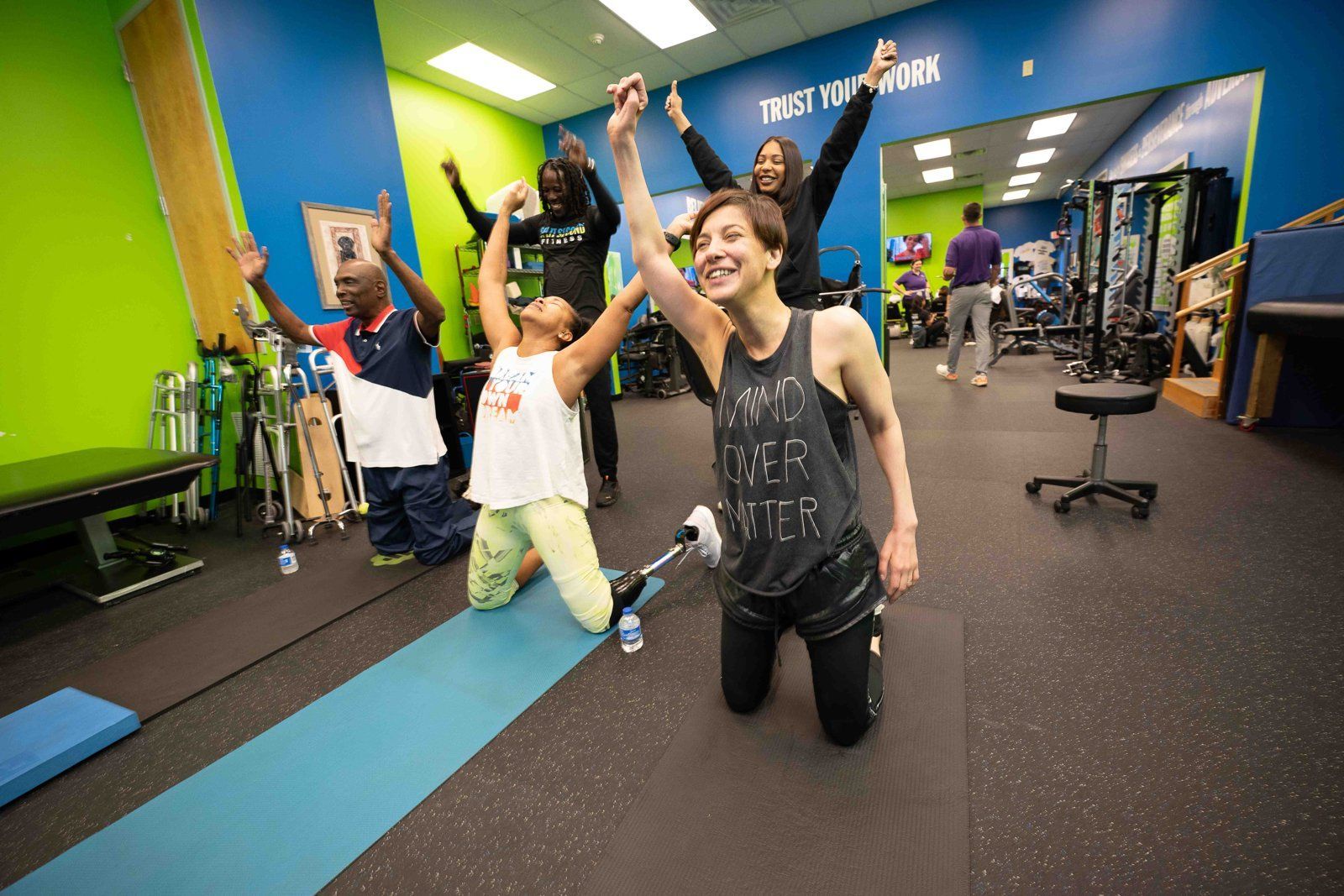 Members kneel on mats and raise their arms in celebration during a group class, one using a prosthetic leg, as two coaches cheer behind them.