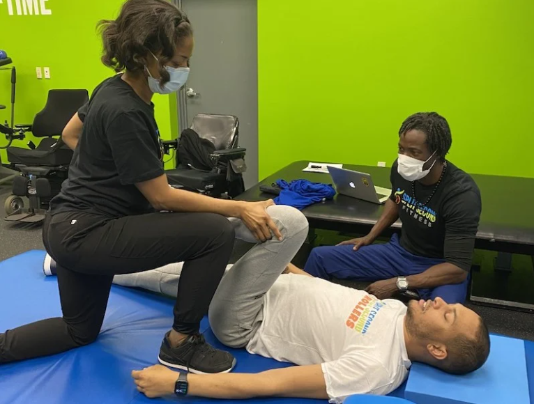 A man is laying on a blue mat while a woman stretches his leg.