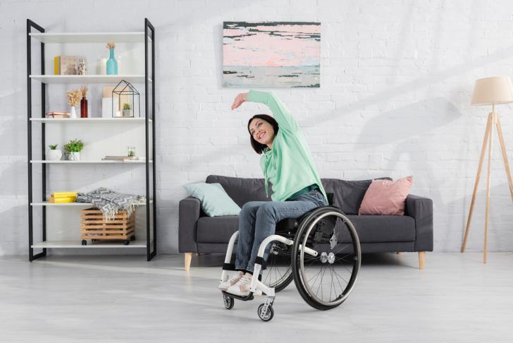 A woman in a wheelchair is stretching her arms in a living room.
