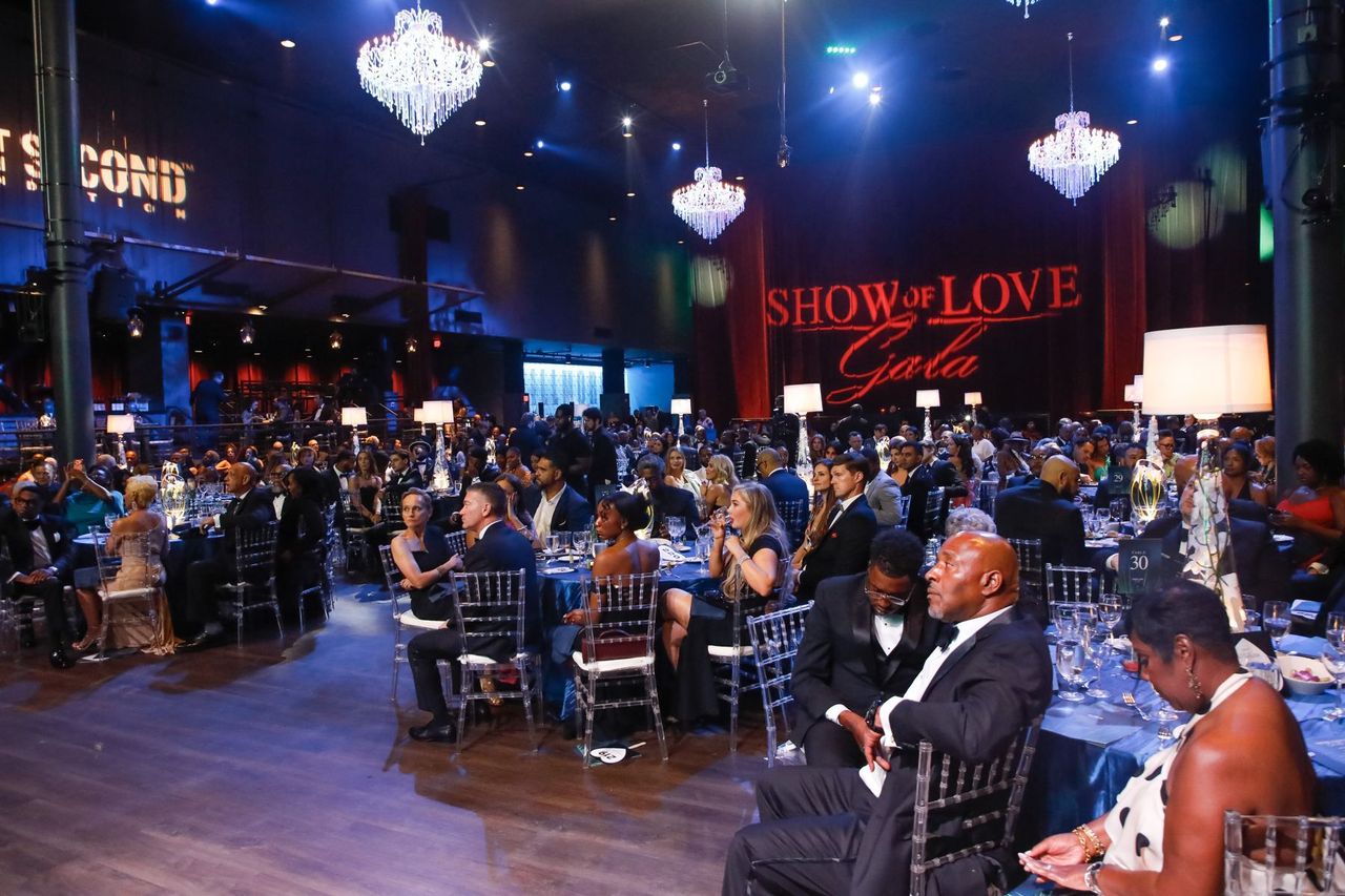 Guests seated at tables beneath chandeliers in the ballroom, with the Show of Love Gala logo projected on the curtain.