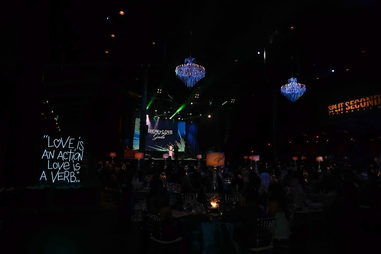 A neon sign reading “Love is an action. Love is a verb.” glows in the darkened ballroom, with the gala stage behind it.