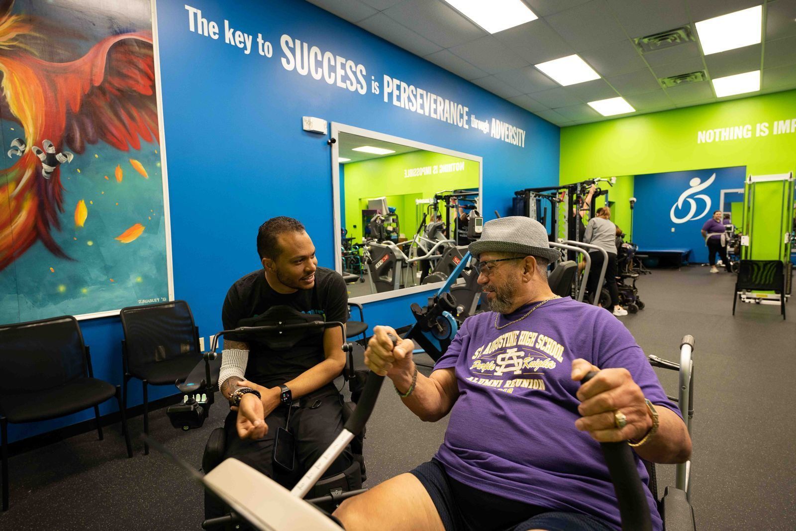 Mark Raymond Jr., in his wheelchair, talks with a member working out on an arm cycle beneath a mural that reads The key to success is perseverance through adversity