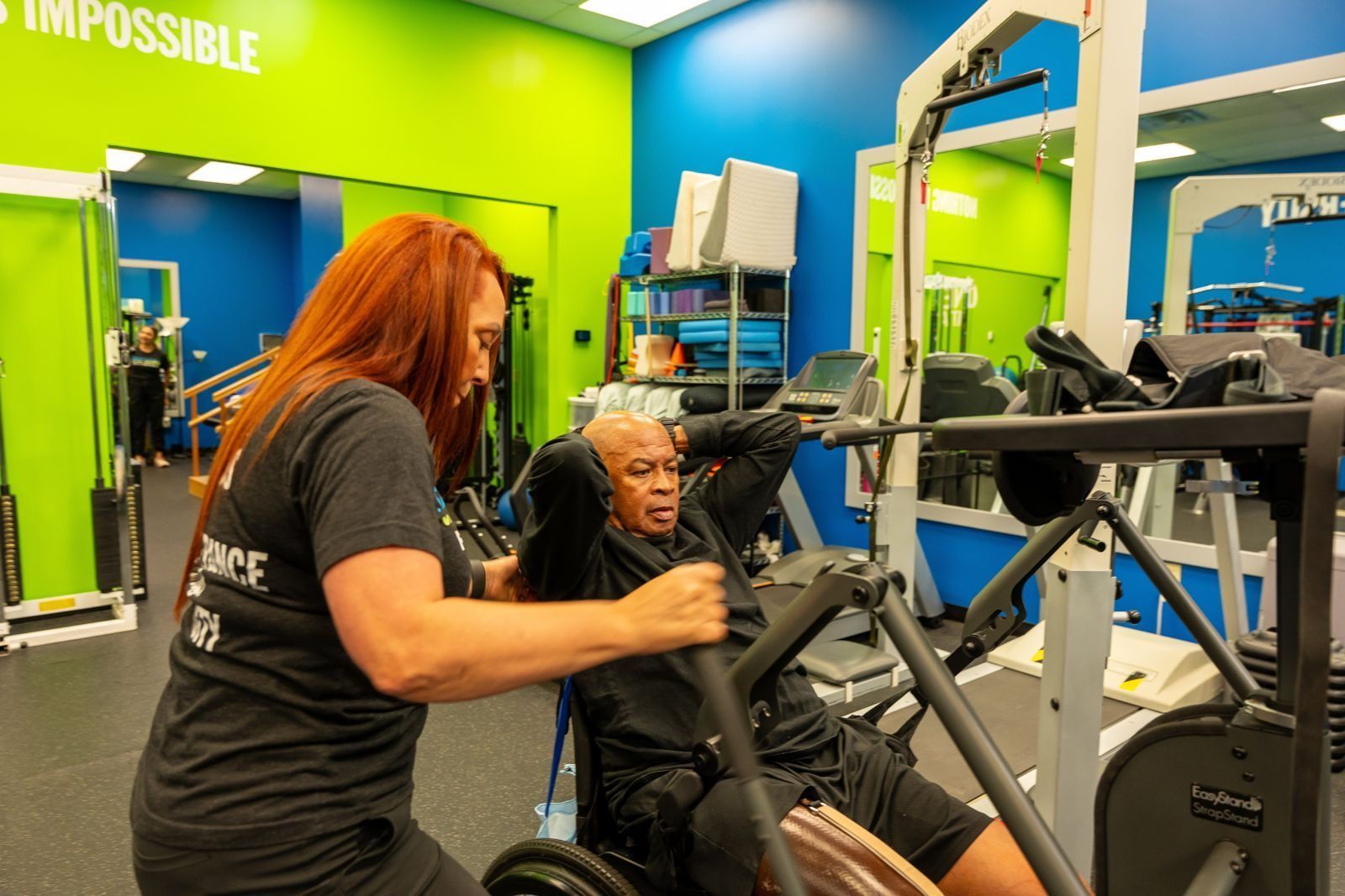 A specialist supports a member through a seated exercise beside a standing frame
