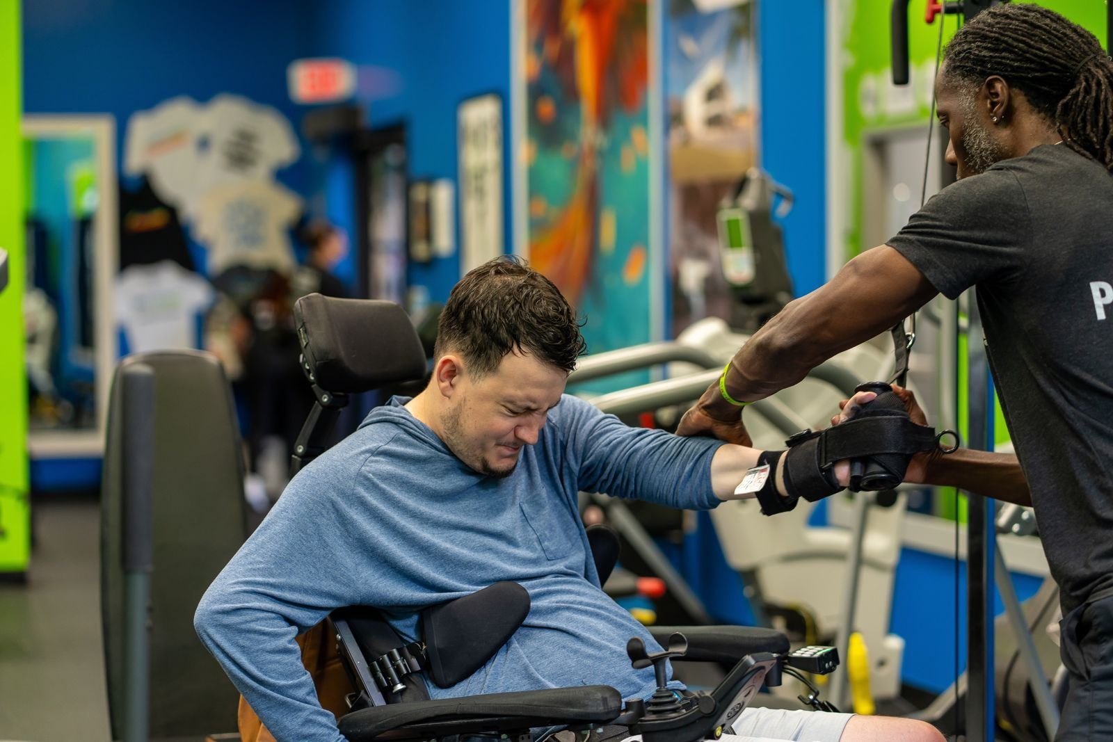 An adaptive fitness specialist guides a member's arm through a cable exercise from his power wheelchair