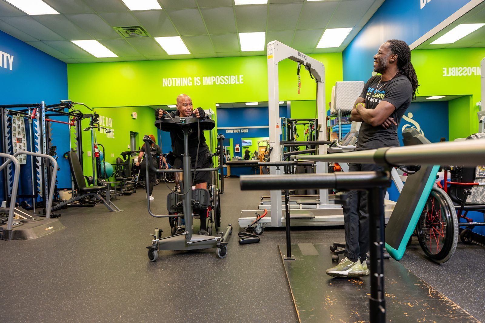 A member uses a standing frame on the training floor while a specialist looks on, under a wall that reads Nothing Is Impossible