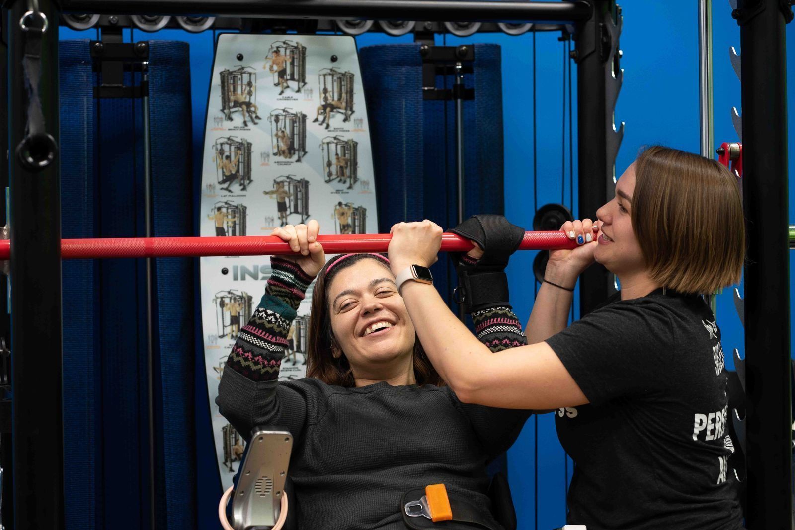 A smiling member in a wheelchair grips a barbell while an adaptive fitness specialist steadies it