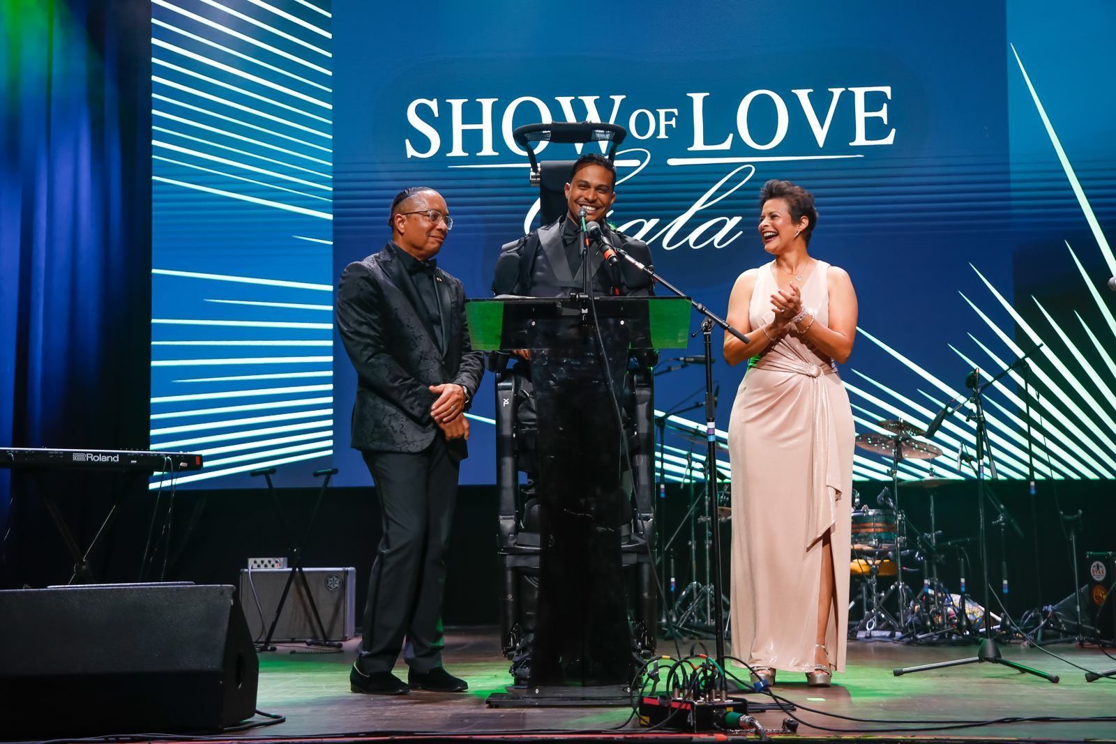 Mark Raymond Jr. speaks from the podium standing in an exoskeleton at the Show of Love Gala, flanked by two guests