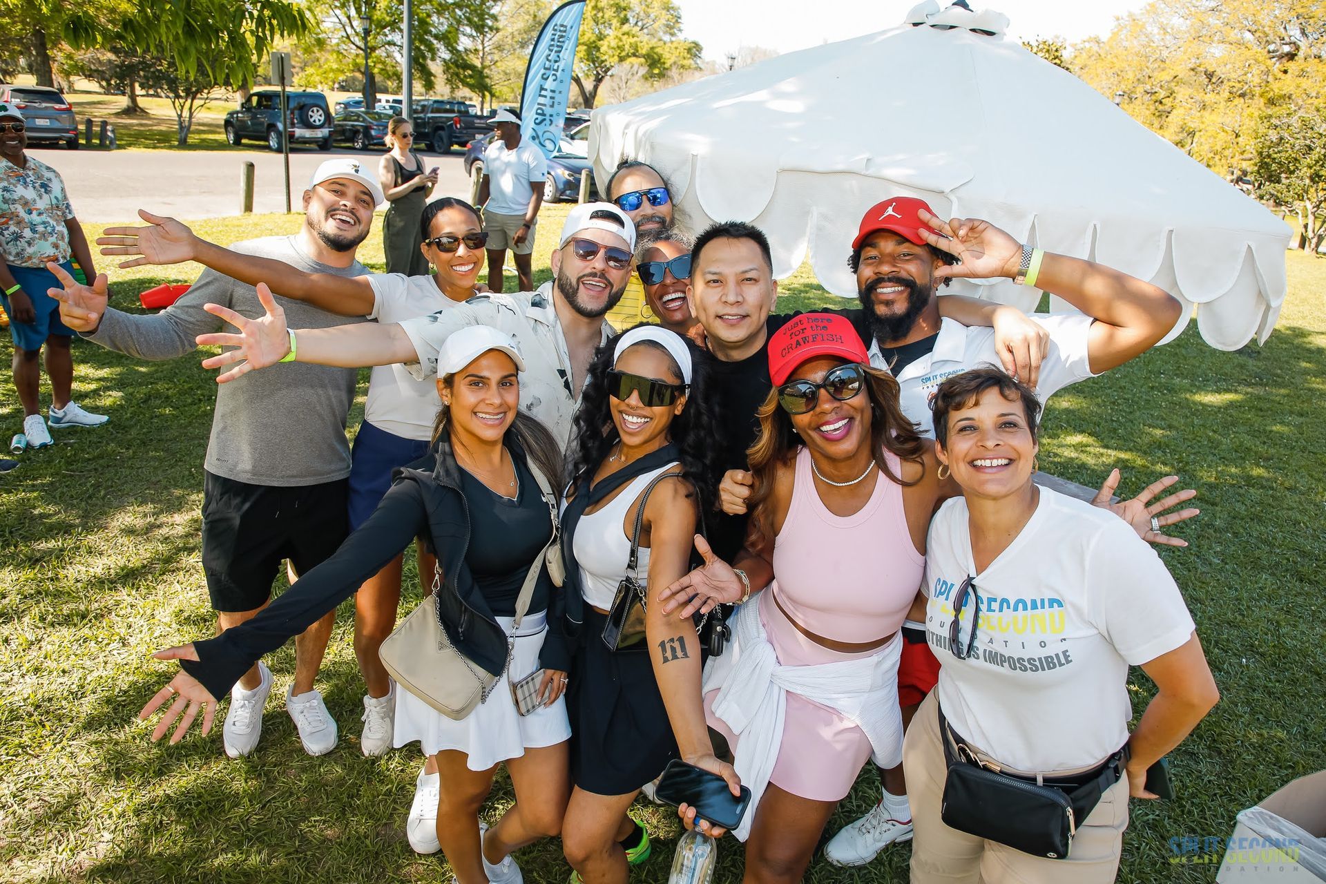 A smiling group of Split Second Foundation friends and supporters, several in Split Second shirts, pose with arms outstretched on the grass at the Spring Picnic.