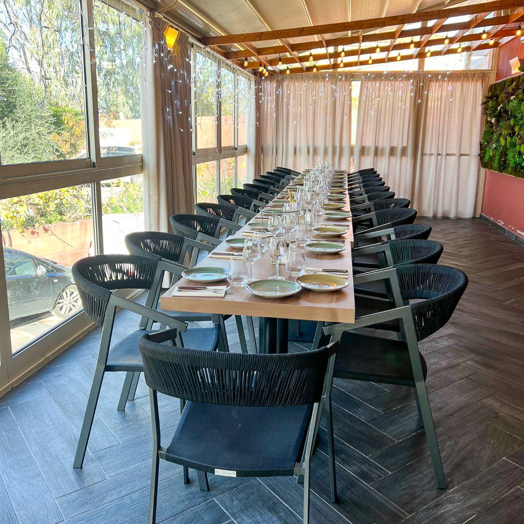 Une longue table à manger est dressée pour un grand groupe dans un patio lumineux et clos, avec un parquet à chevrons foncé et des chaises noires.