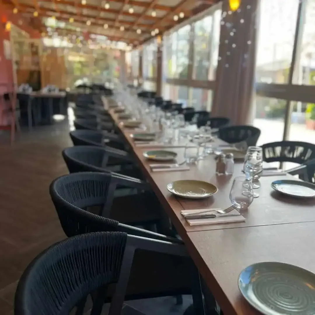 Une longue table à manger en bois dressée pour un événement formel dans un restaurant, avec des chaises sombres et des guirlandes lumineuses au plafond.