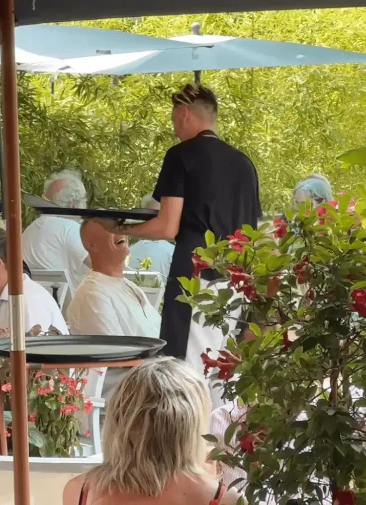 Un serveur tient un plateau au-dessus de la tête d'un client hilare sur la terrasse d'un restaurant en plein air, agrémentée de parasols et de verdure.