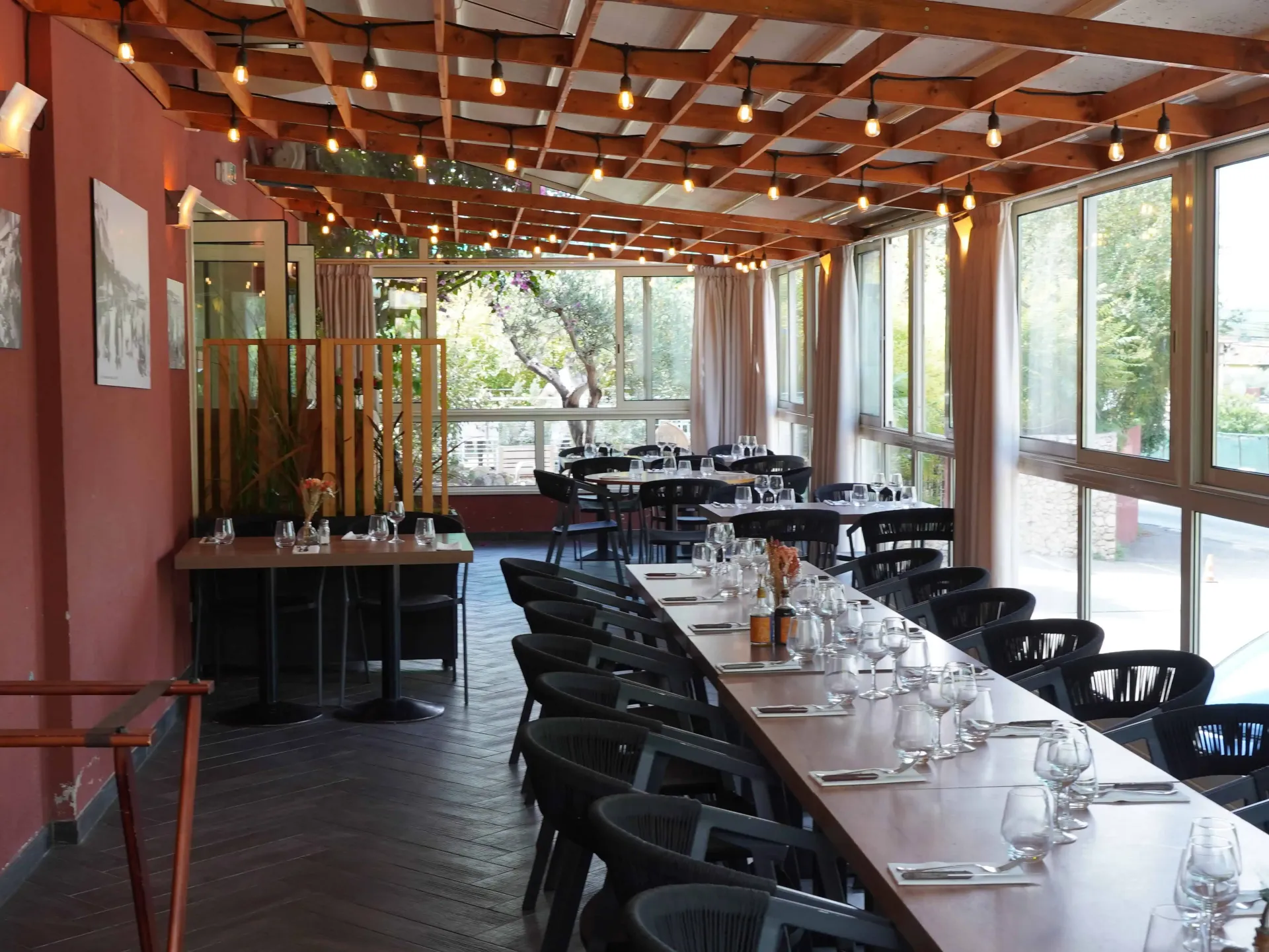 Un coin repas comprenant une longue table en bois, des chaises noires, un plafond en bois à lattes avec des suspensions lumineuses et de grandes fenêtres.