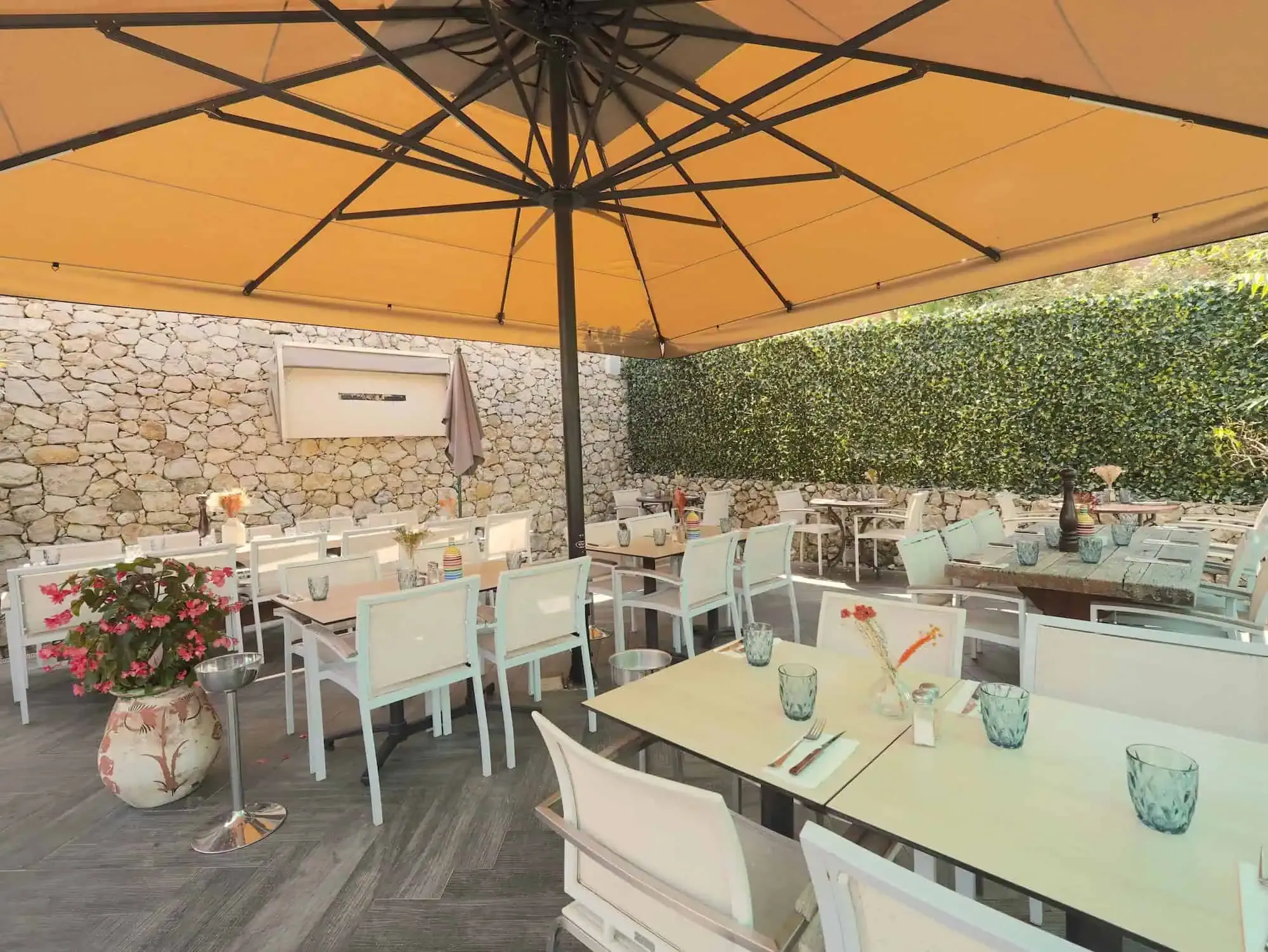 Terrasse extérieure de restaurant avec tables, chaises blanches et un grand parasol beige sous un mur de pierre et une haie de lierre.