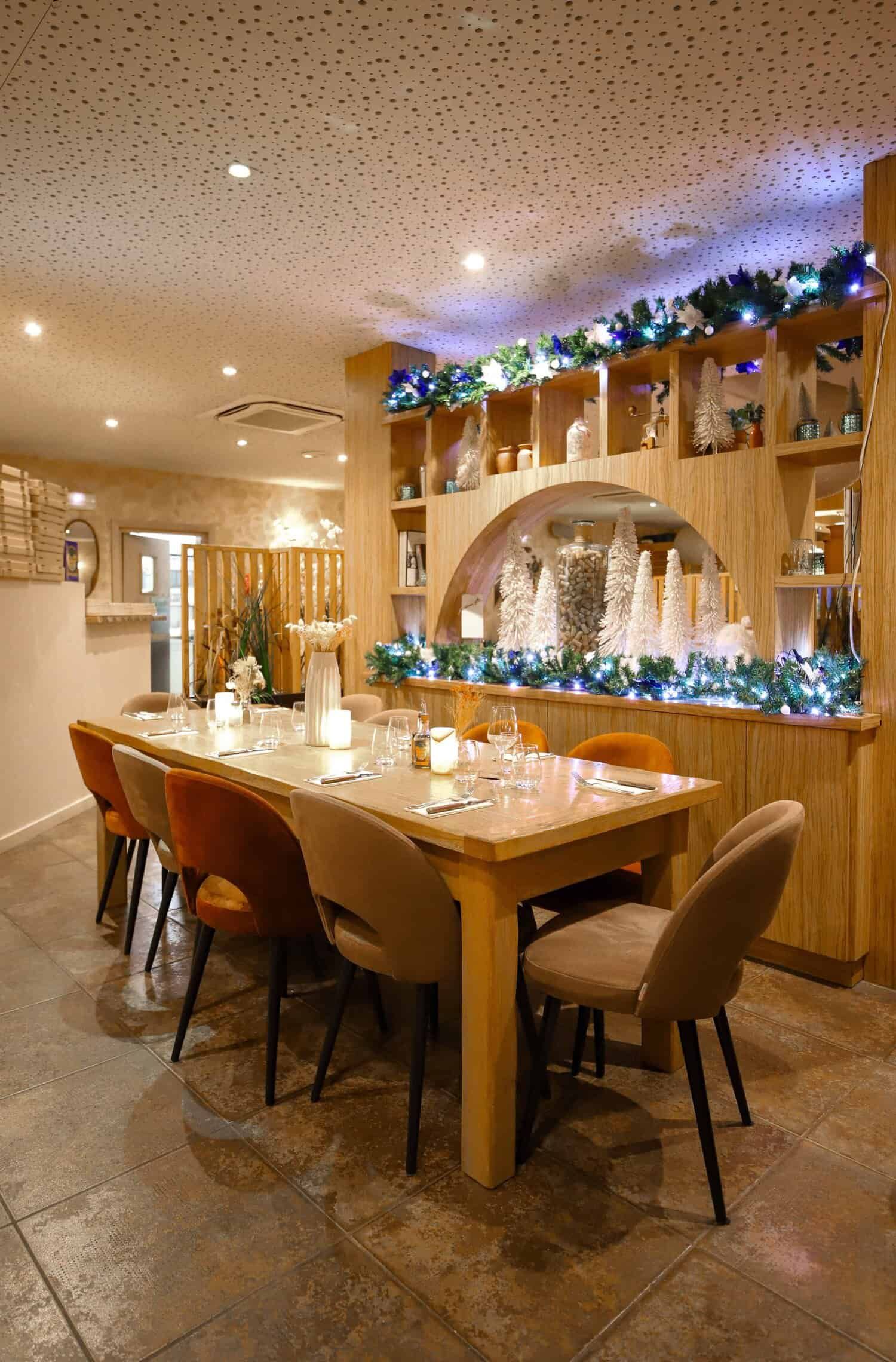 Une table à manger dressée pour un groupe dans une pièce festive aux tons chauds, avec des étagères en bois décorées de guirlandes lumineuses.