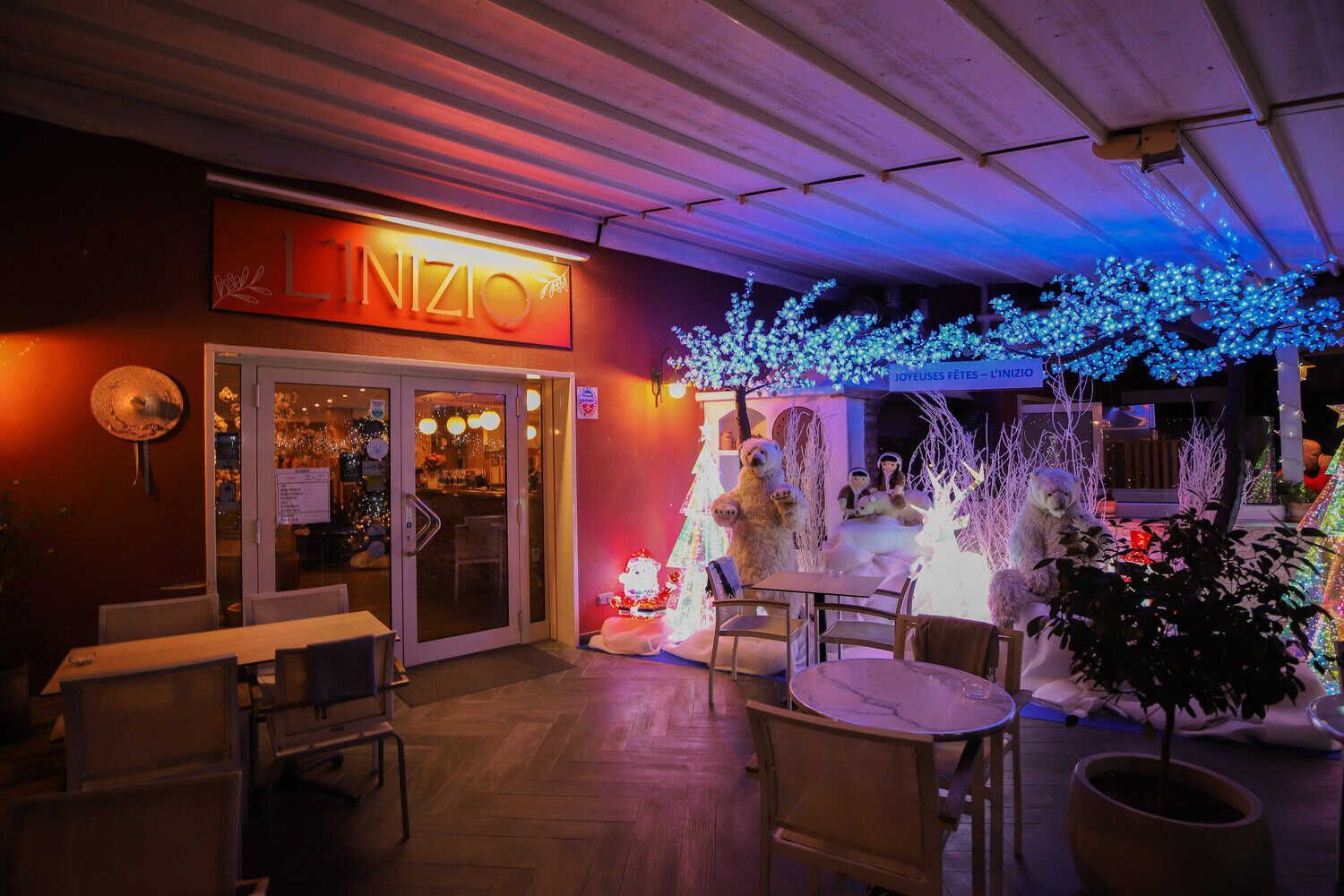 Vue extérieure de l'entrée d'un restaurant la nuit, illuminée par des guirlandes lumineuses bleues et des lampes à lumière chaude, avec une terrasse.