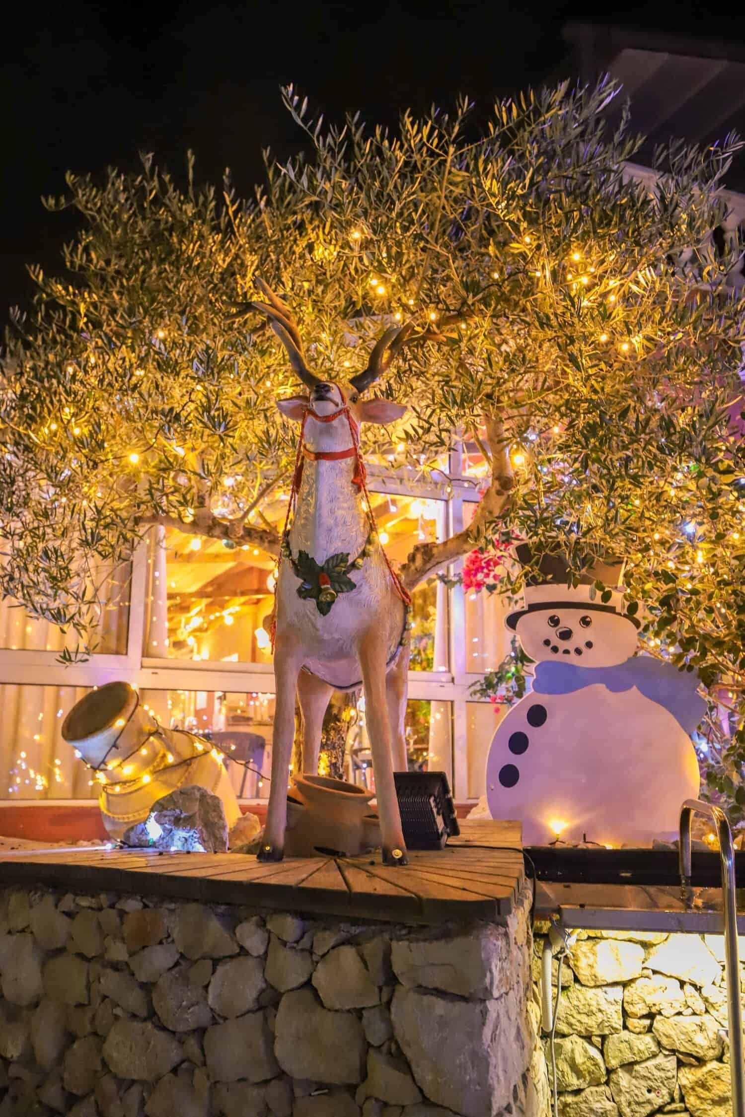 La nuit, un arbre illuminé, décoré de guirlandes lumineuses, se dresse à côté d'une grande statue de renne et d'un bonhomme de neige souriant.