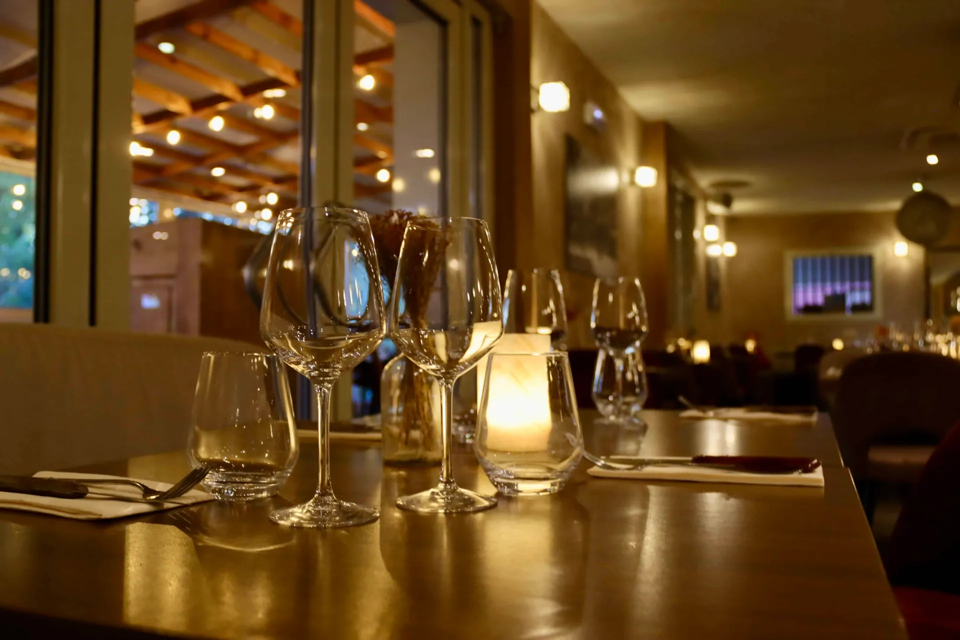 Une table de restaurant dressée avec des verres à vin et une bougie allumée, dans une ambiance intérieure chaleureuse et tamisée.