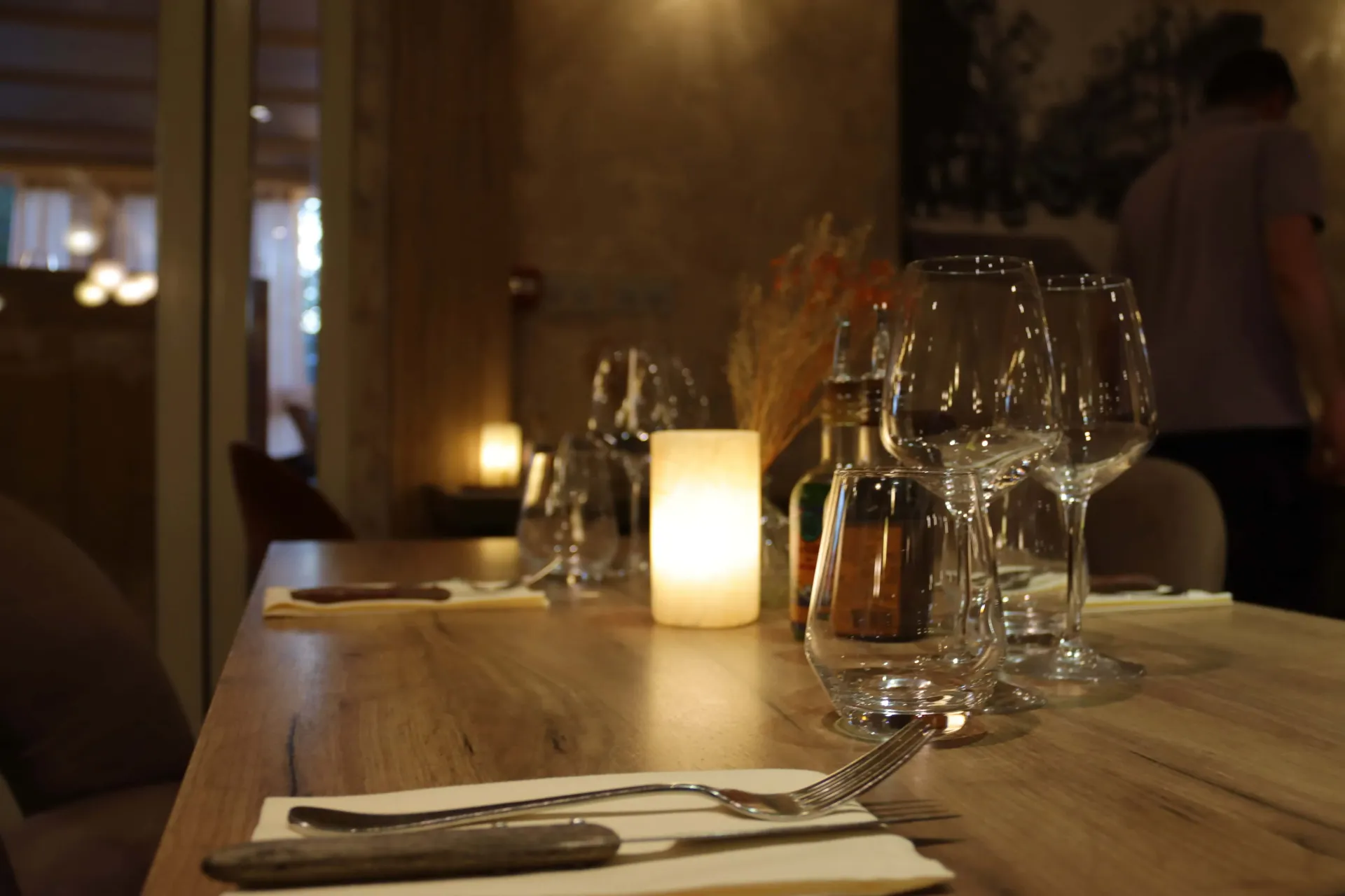 Une table de restaurant chaleureuse et tamisée, dressée avec des verres à vin, une bougie qui brille et des couverts.