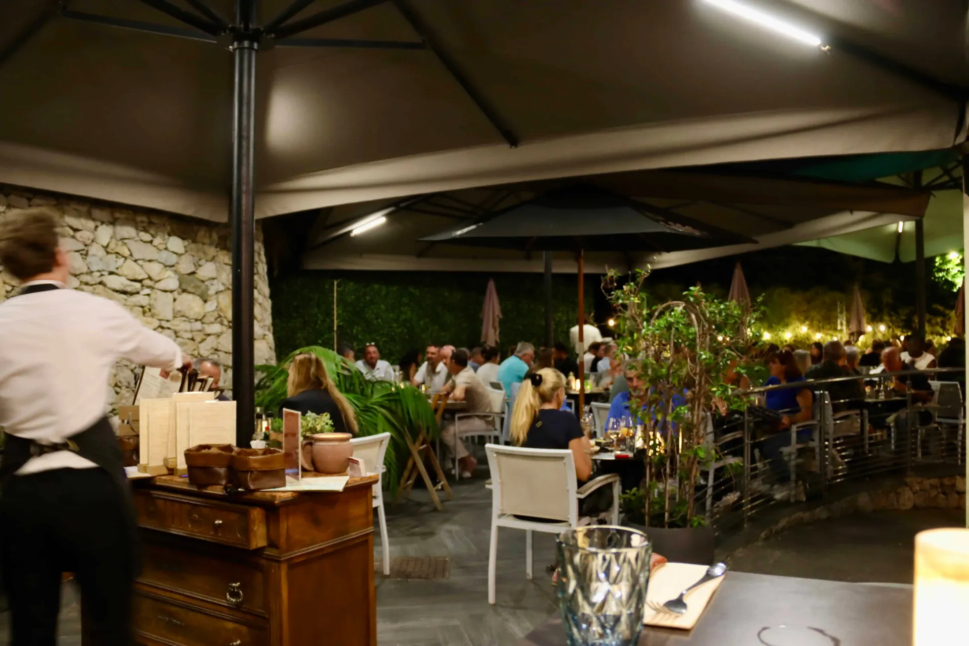 Une terrasse de restaurant en plein air le soir, avec des clients assis à des tables sous de grands parasols et un membre du personnel à proximité.
