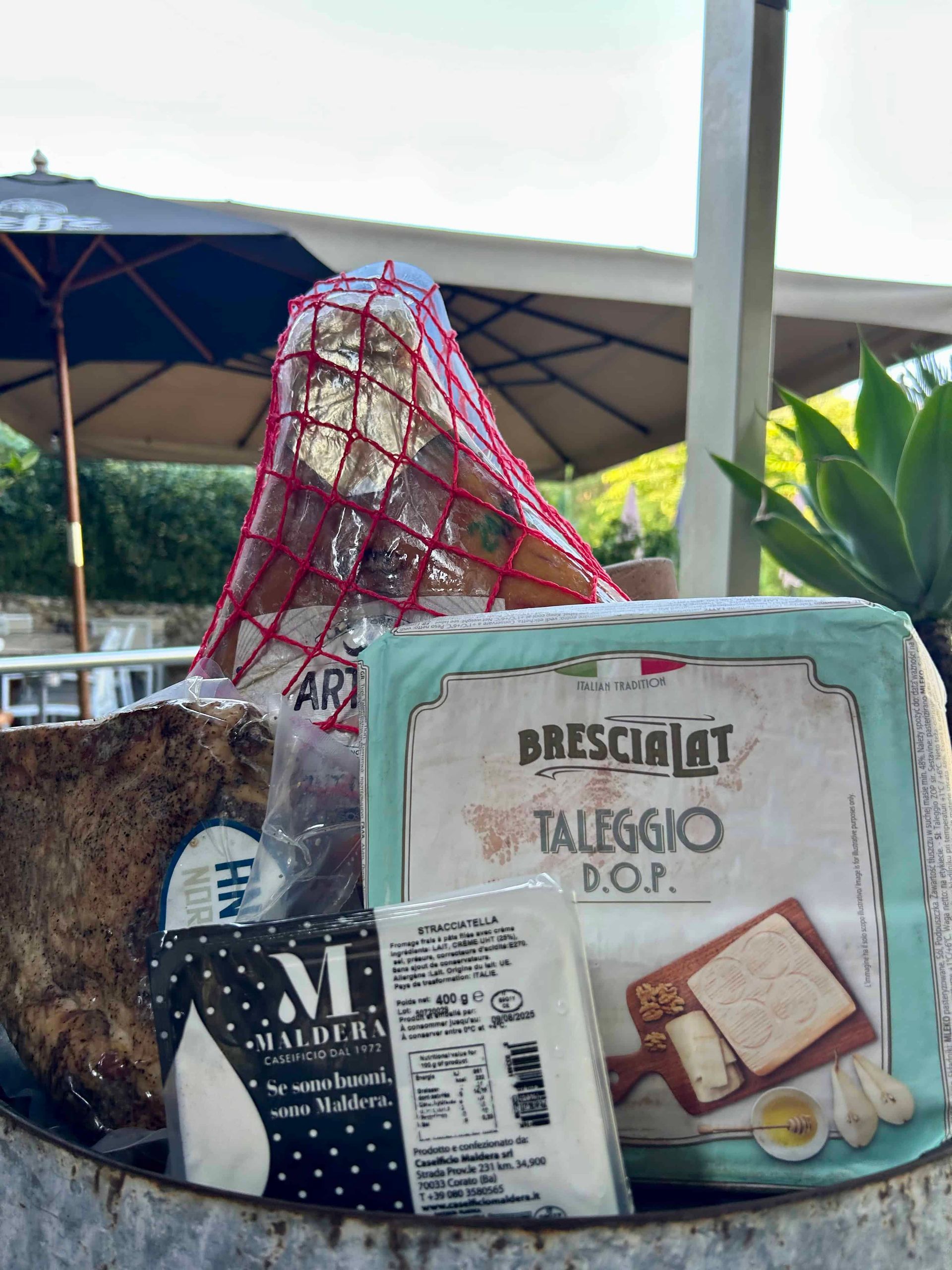 Un panier-cadeau contenant du fromage Brescialat Taleggio, des produits laitiers de Maldura et de la charcuterie emballée dans un filet, disposé sur une terrasse.