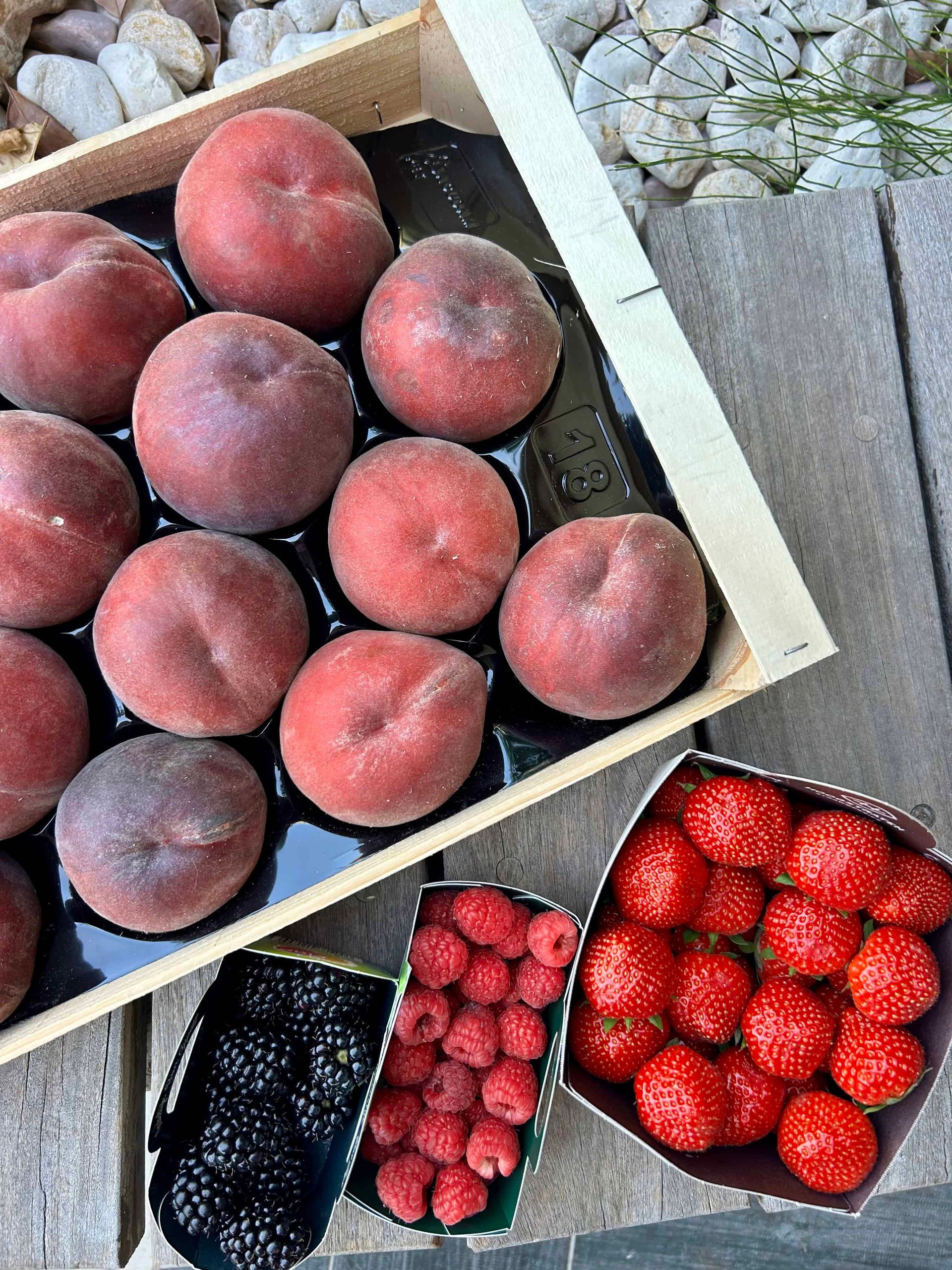 Une caisse en bois remplie de pêches, posée à côté de récipients contenant des mûres, des framboises et des fraises, sur une surface en bois.