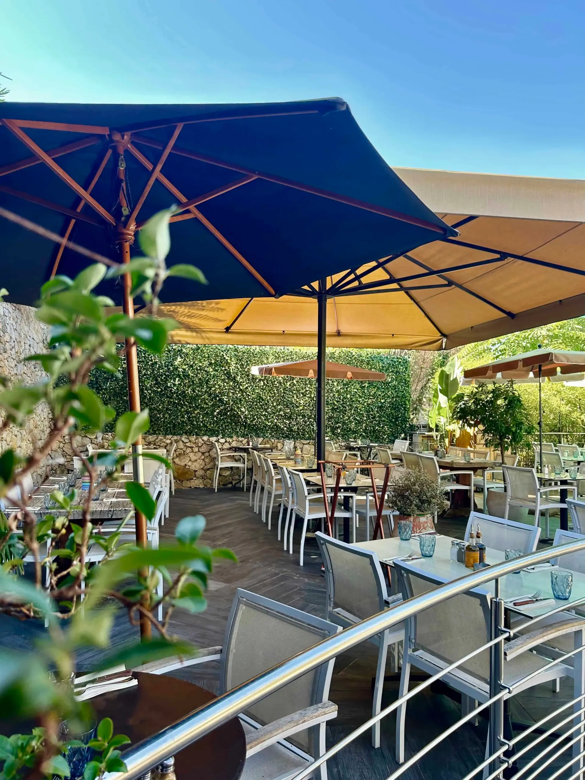 Une terrasse de restaurant en plein air avec des tables, des chaises et de grands parasols bleus et beiges sous un ciel ensoleillé.