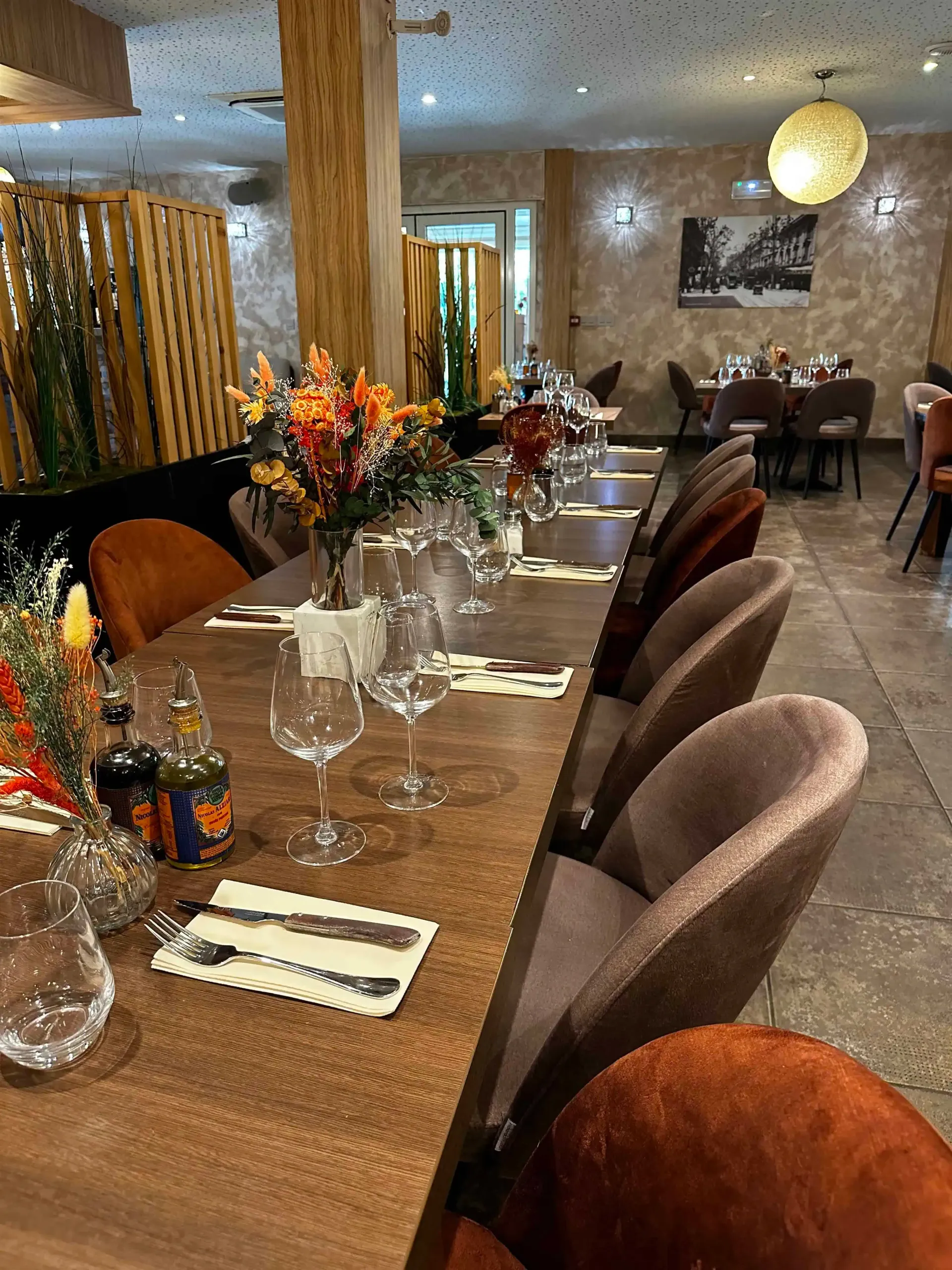 Dans un restaurant, une longue table est dressée pour un repas, avec de la verrerie, des couverts et des centres de table floraux.