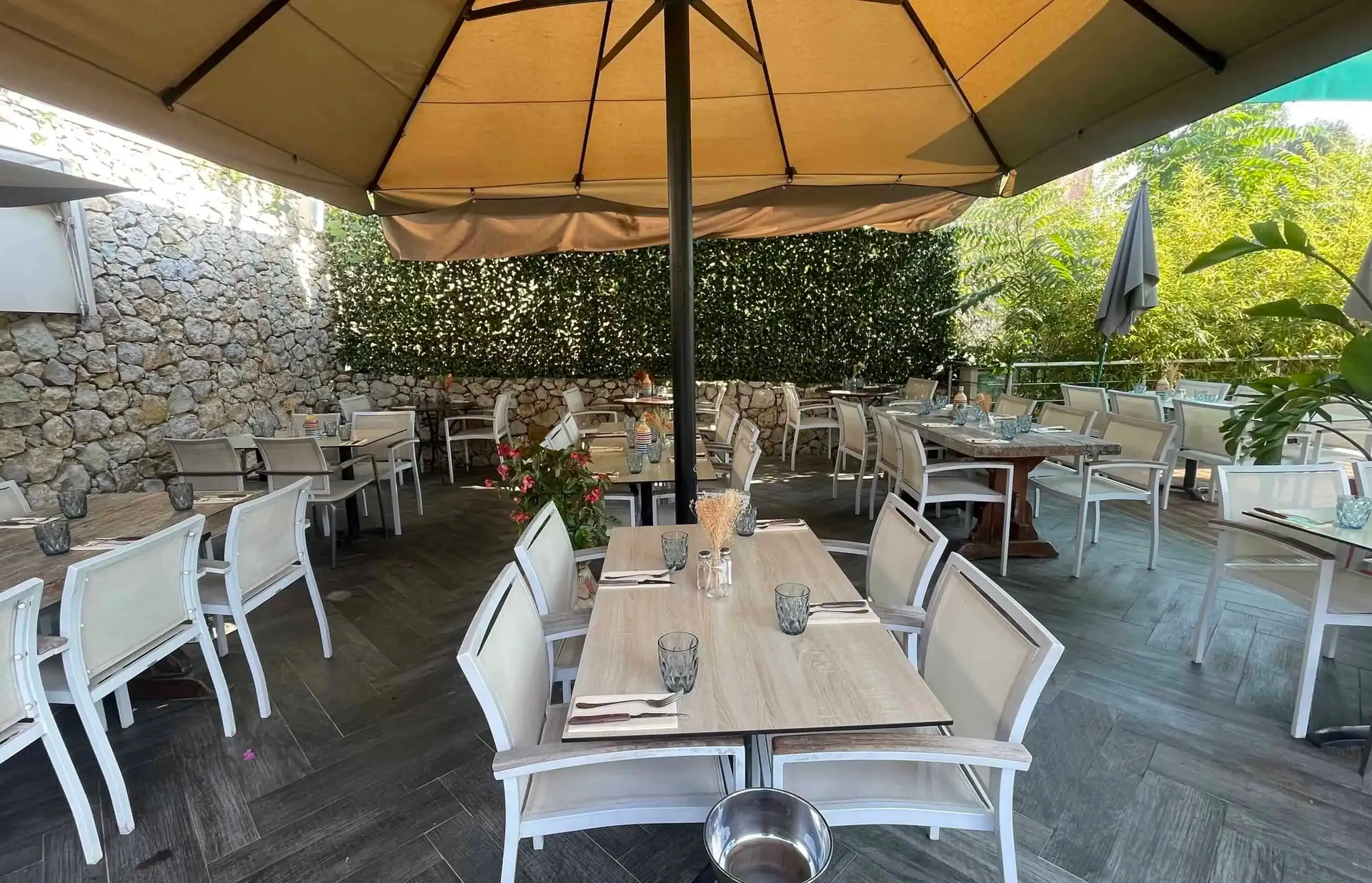 Terrasse extérieure de restaurant avec tables et chaises vides, et un grand parasol beige sous un muret en pierre.