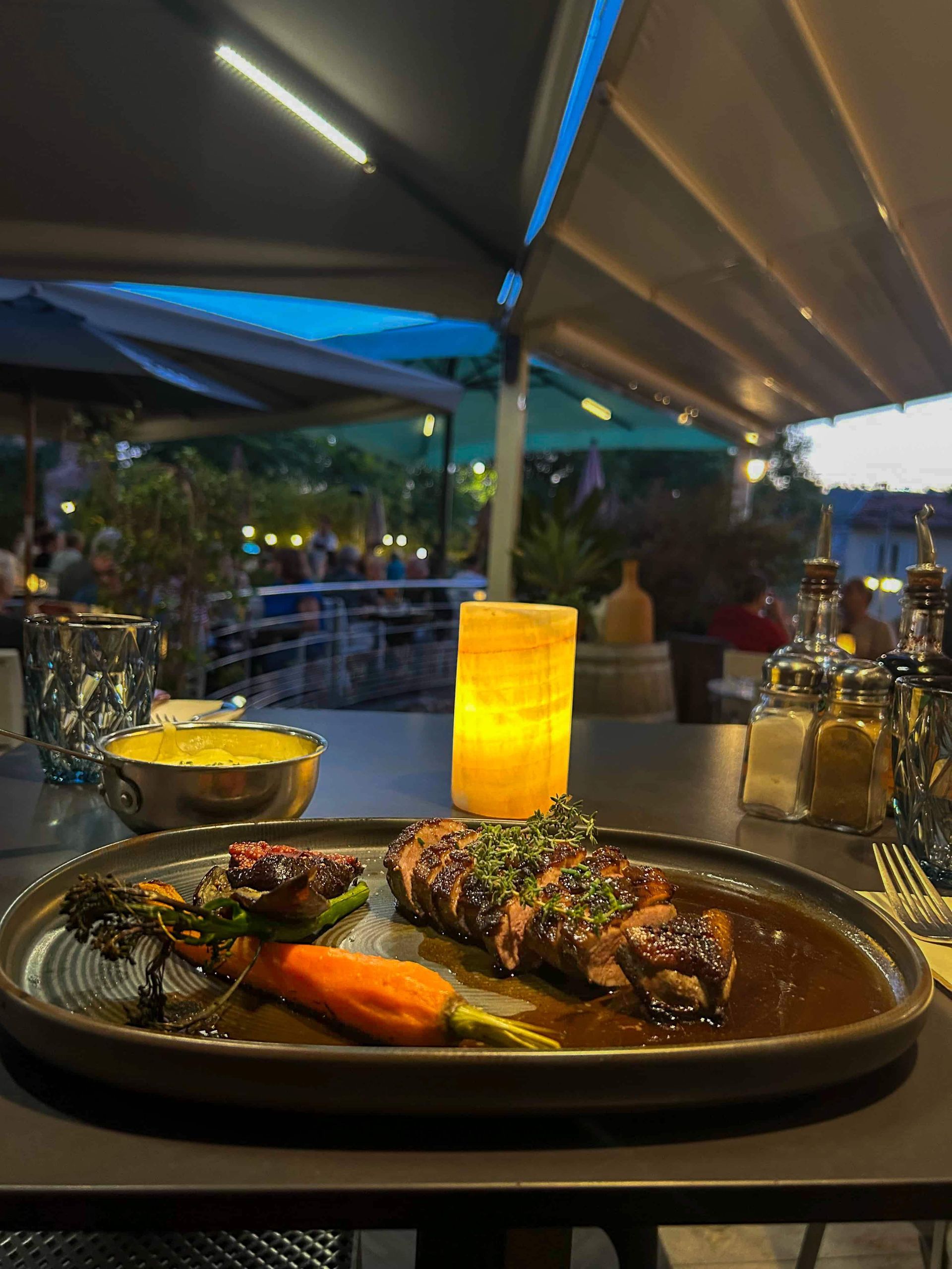 Un dîner gastronomique servi à l'assiette, composé de viande rôtie, de carottes glacées et d'herbes aromatiques, sur une terrasse extérieure au crépuscule.