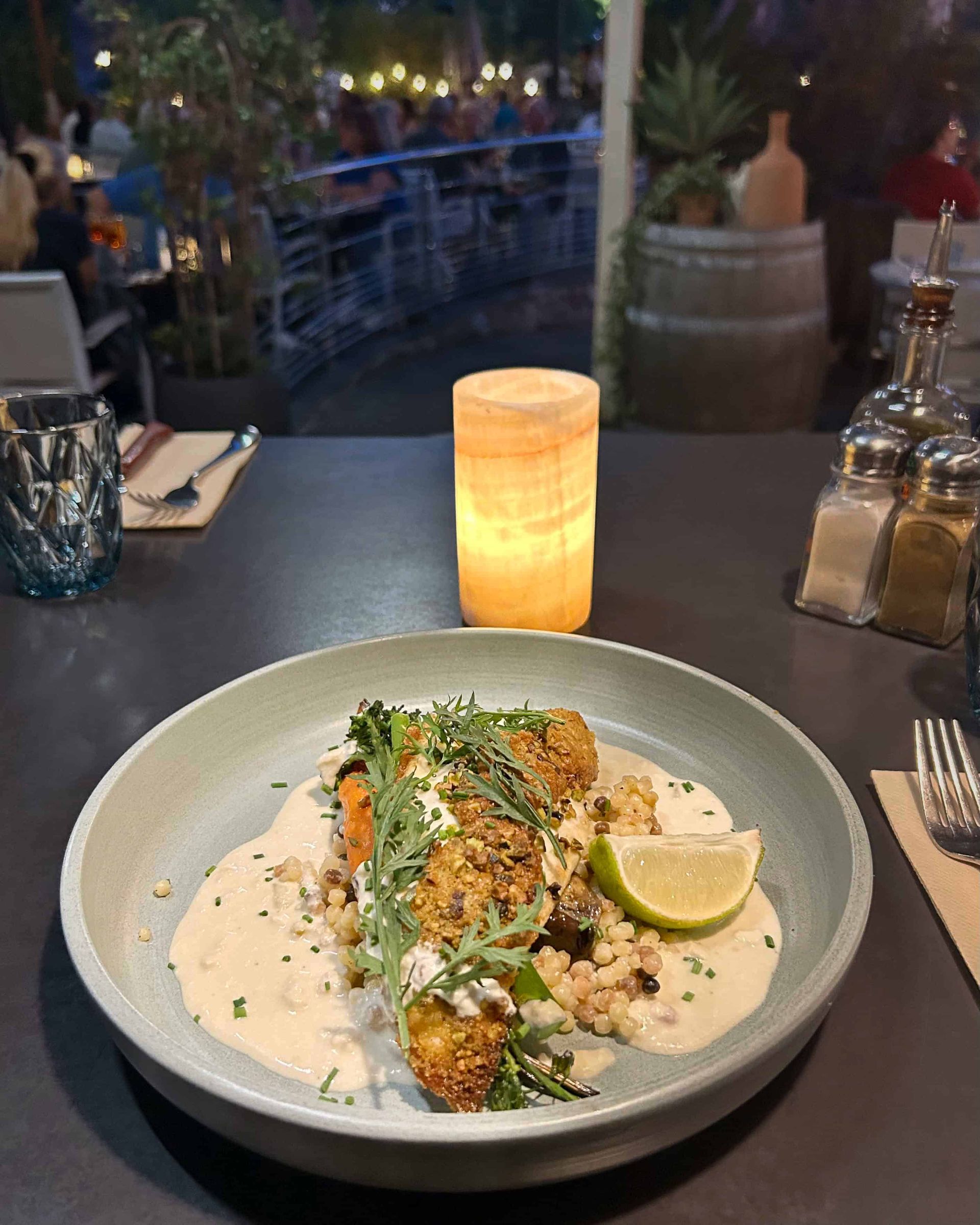 Une assiette de poisson pané avec une sauce crémeuse, un quartier de citron vert et de la verdure, servie dans un restaurant en plein air à la lueur des bougies.