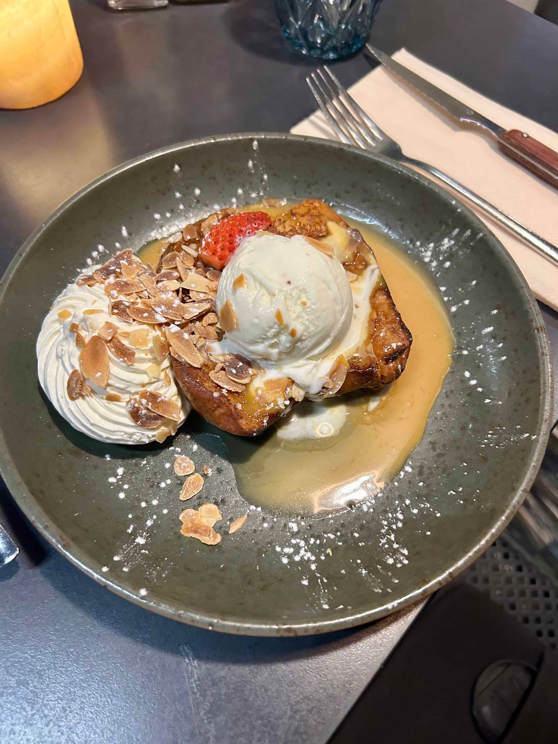 Une tranche de pain perdu garnie d'une boule de glace à la vanille, de crème fouettée, d'amandes effilées et de sirop, servie dans une assiette.