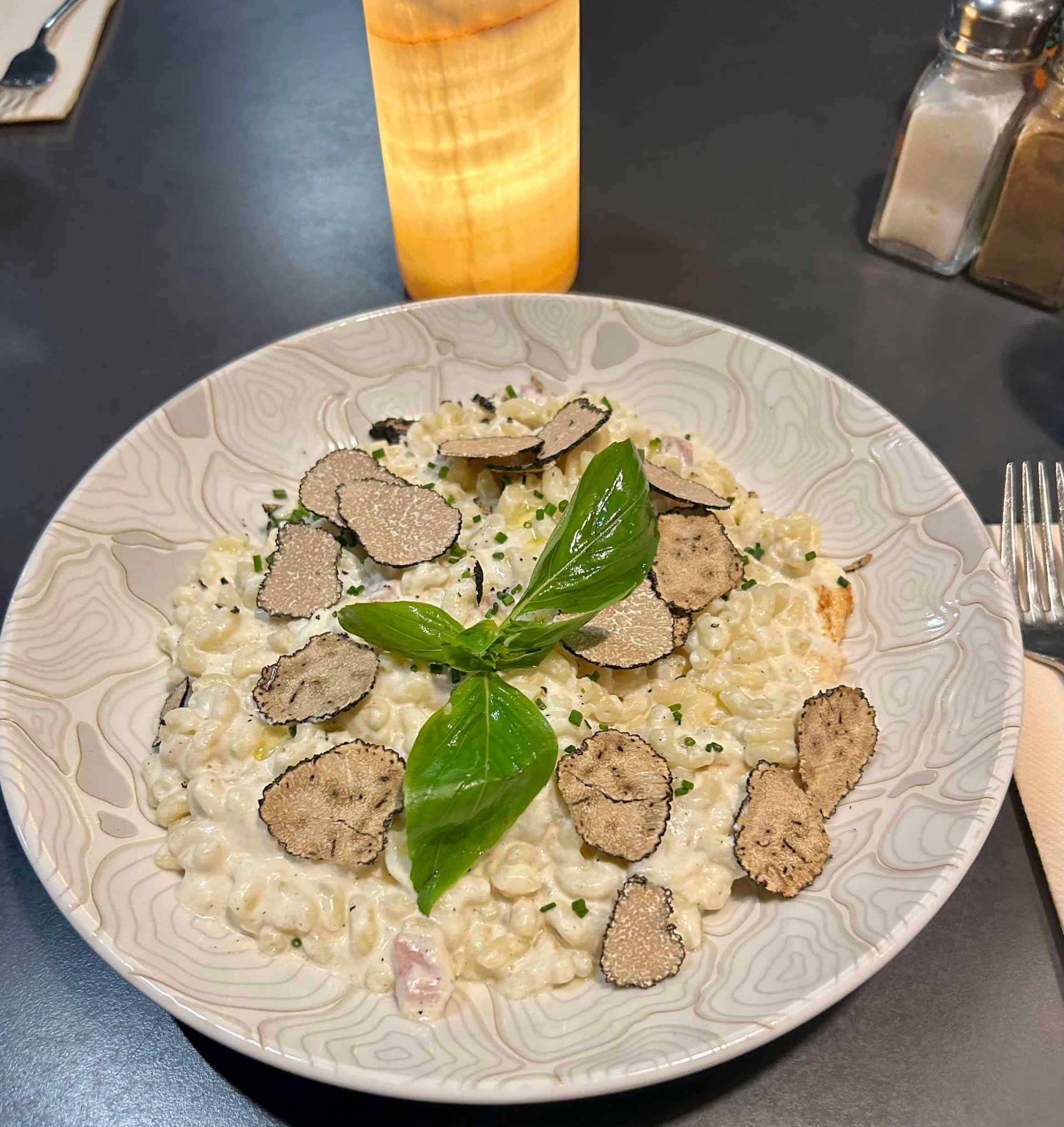 Une assiette de pâtes crémeuses garnie de tranches de truffe fraîche et d'une feuille de basilic, posée sur une table sombre à côté d'une lampe.