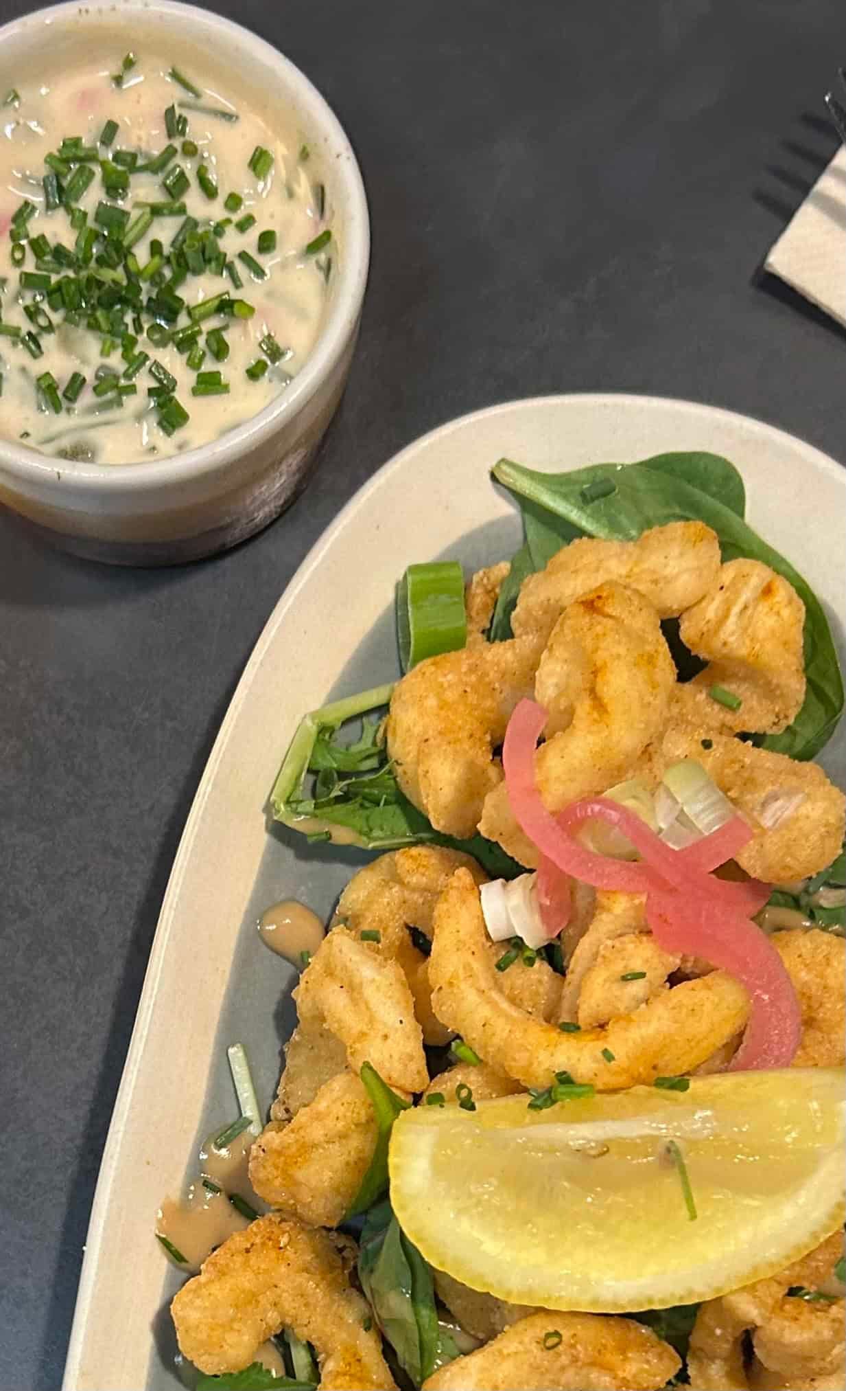 Une assiette de calamars frits avec des légumes verts, des oignons marinés et un quartier de citron, accompagnée d'un petit bol de sauce crémeuse.