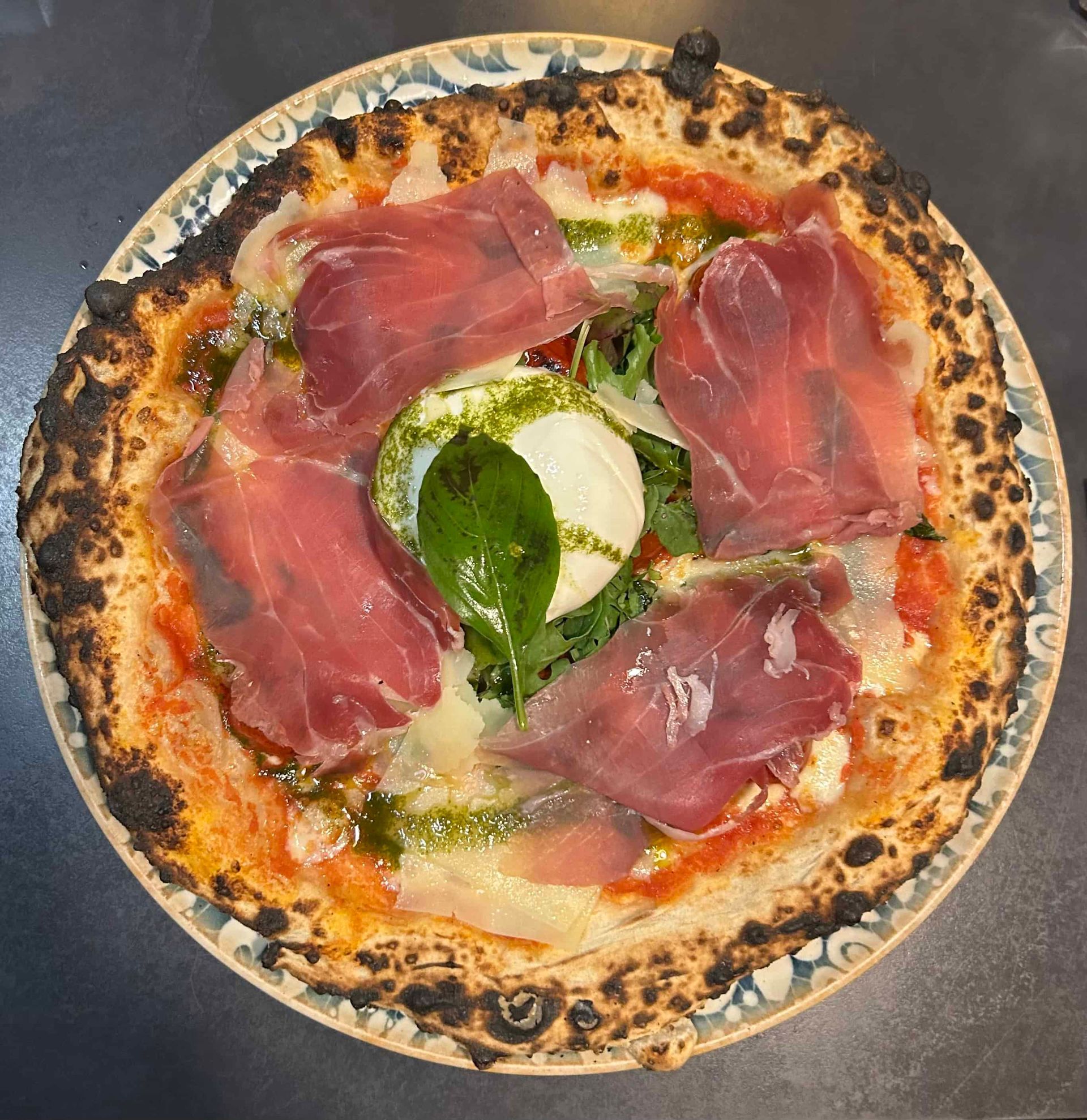 Une pizza napolitaine avec sauce tomate, tranches de prosciutto, pesto, copeaux de fromage et une feuille de basilic frais sur le dessus.