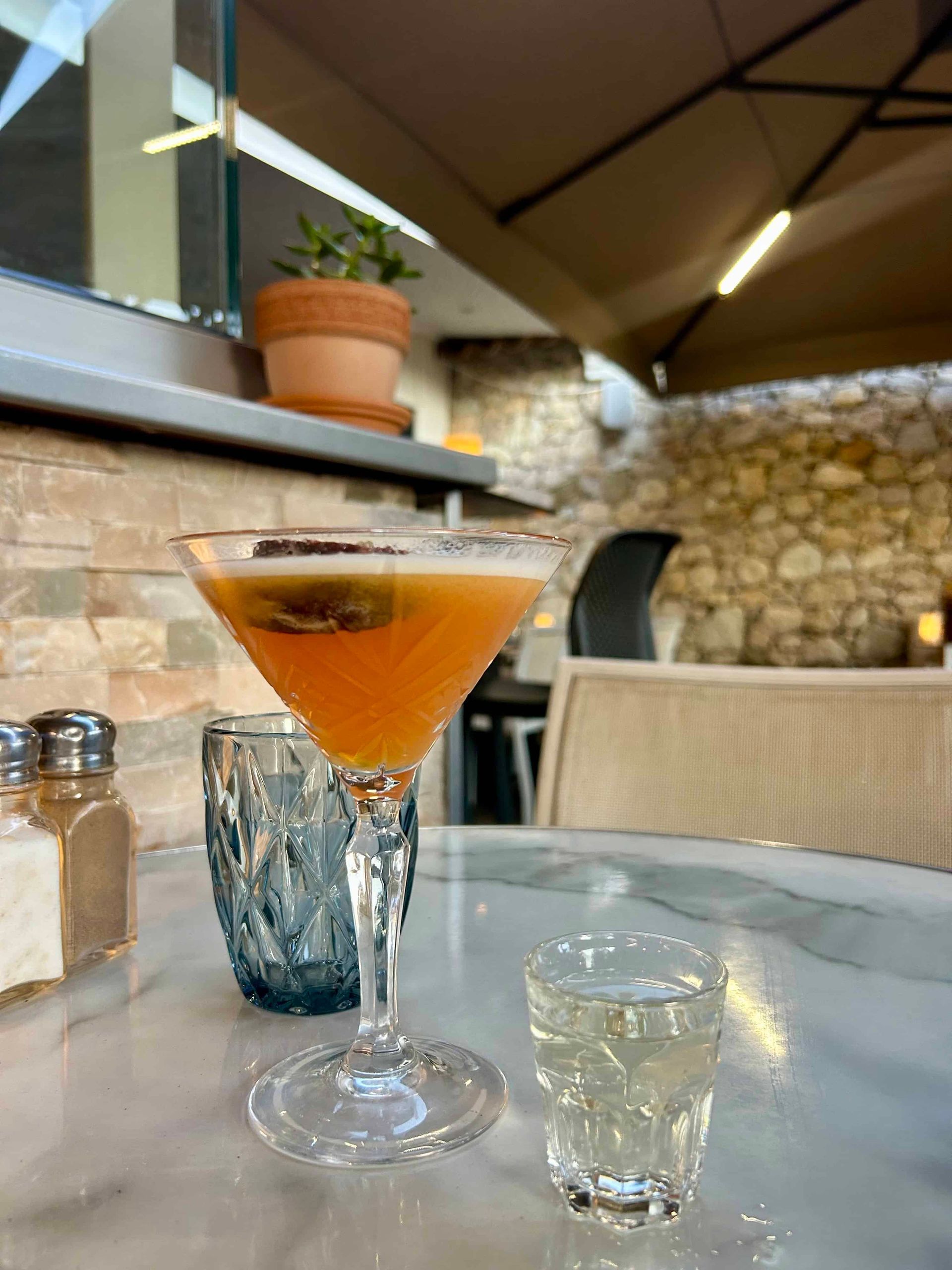 Un cocktail à l'orange servi dans un verre à pied repose sur une table en marbre, à côté d'un petit verre à liqueur transparent et d'un gobelet bleu à motifs.