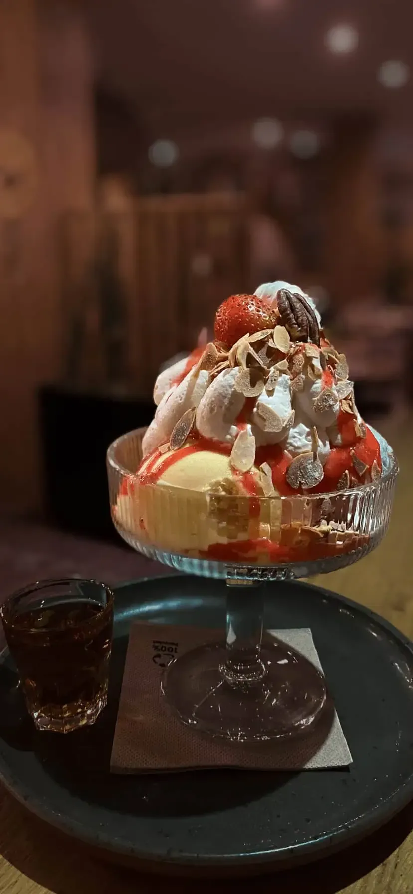 Une coupe glacée composée de glace, de crème fouettée, de fruits rouges et de sauce, servie dans un verre sur un plateau et accompagnée d'un filet de sirop foncé.