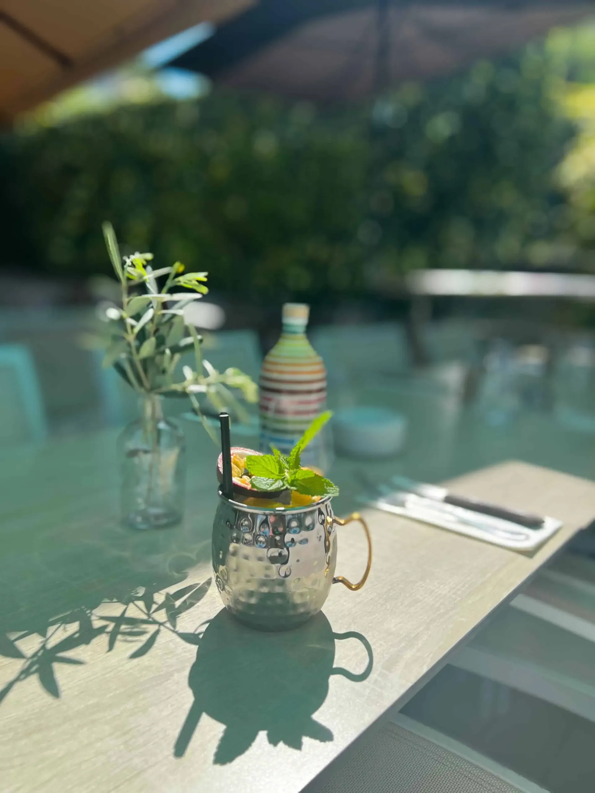 Un cocktail servi dans une tasse en métal avec une garniture de menthe, une plante feuillue dans un bocal en verre et des couverts sur une table de terrasse.