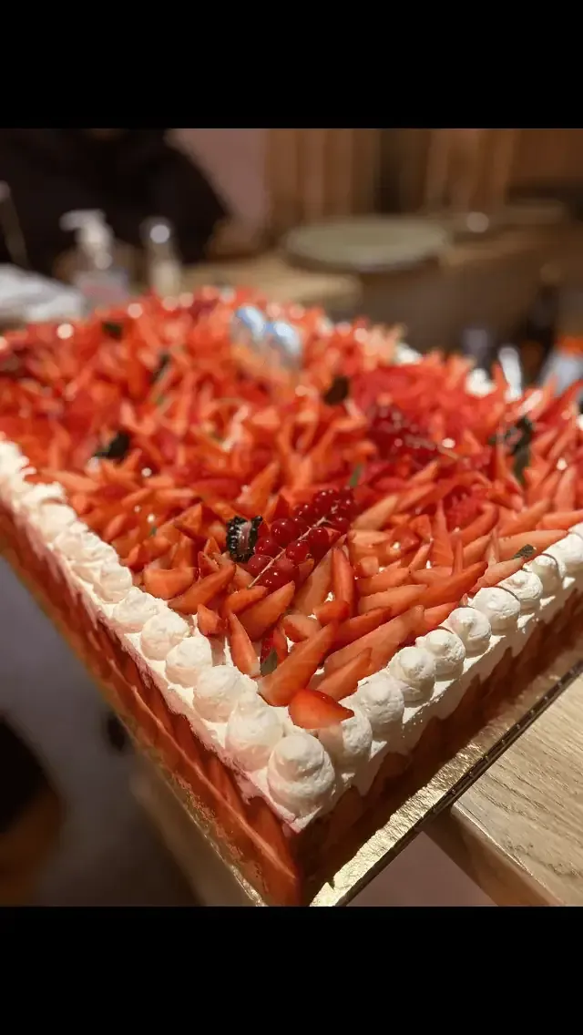 Un gâteau rectangulaire garni de nombreuses tranches de fraises et d'une bordure de crème blanche dressée à la poche à douille, posé sur une table en bois.