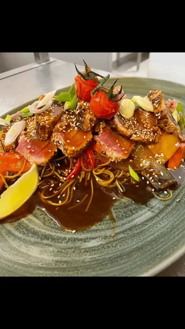 Une assiette de thon saisi garnie de tomates cerises, de graines de sésame et d'oignons verts, servie sur des nouilles avec une sauce foncée.