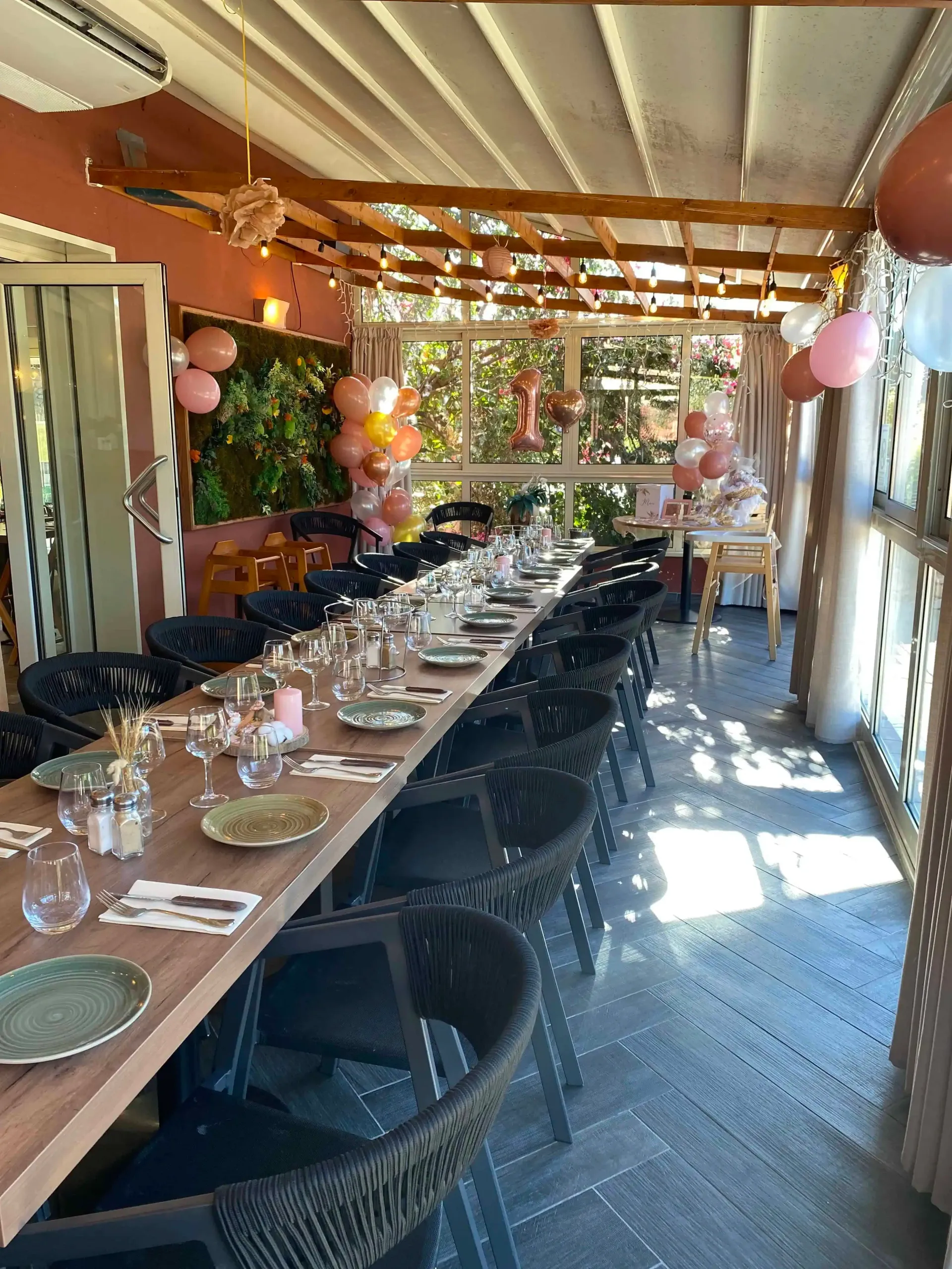 Une longue table à manger en bois dressée pour une fête, décorée de ballons, dans un patio extérieur couvert et ensoleillé.