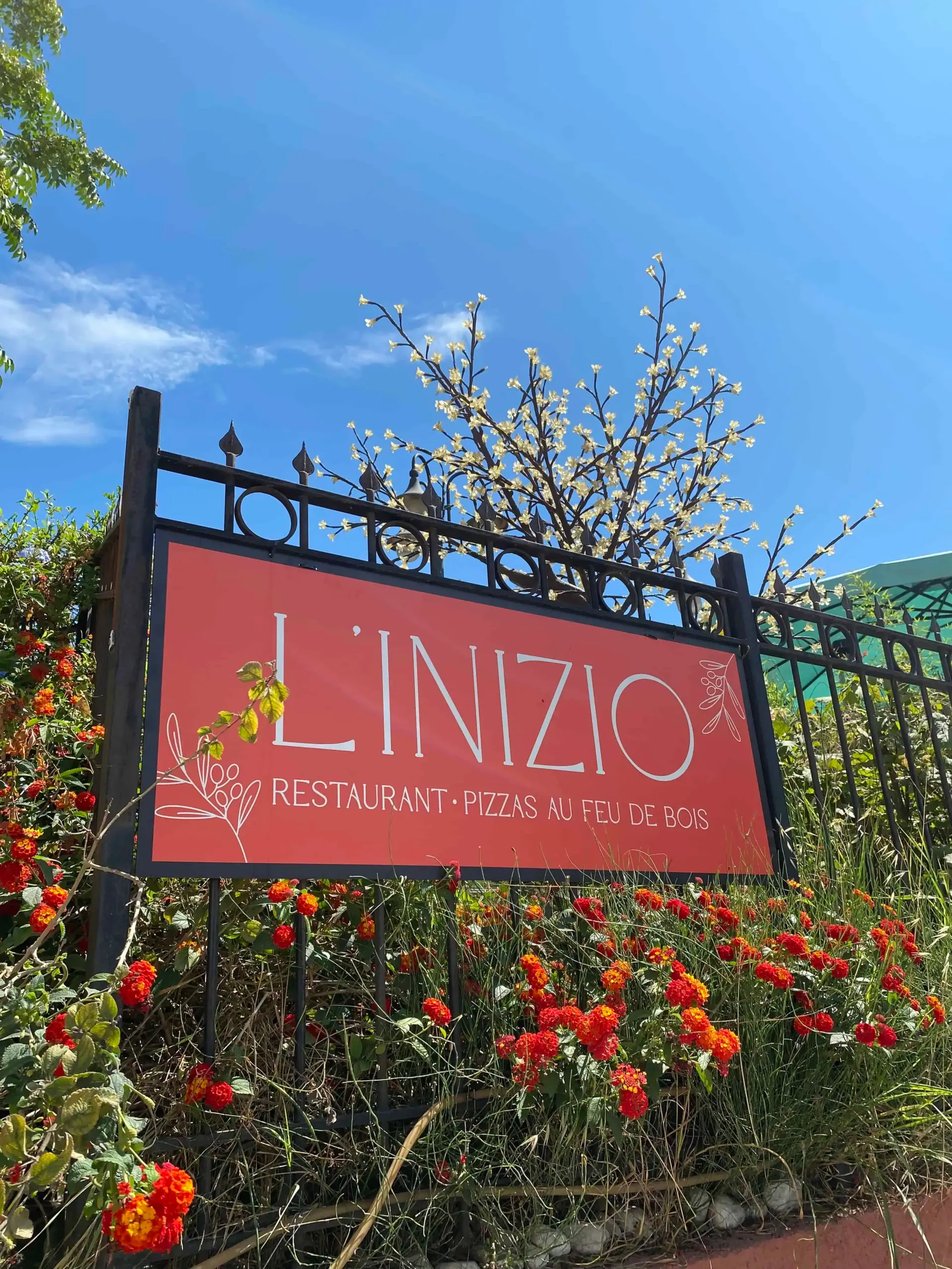 Une pancarte rouge portant l'inscription « L'INIZIO » entourée de roses rouges et de buissons fleuris se détachant sur un ciel d'un bleu éclatant.