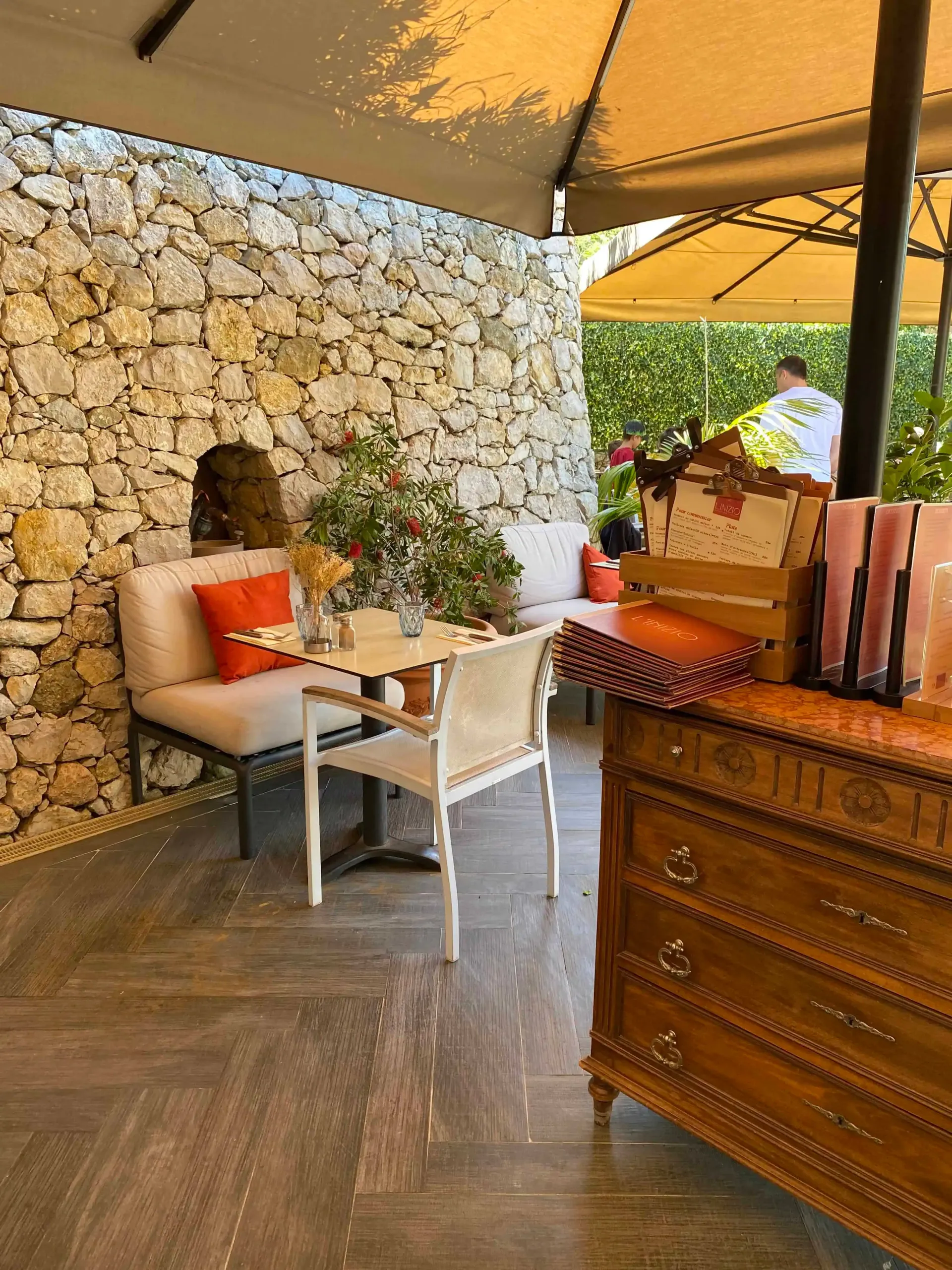 Un coin salon sur la terrasse avec un muret en pierre, une petite table, une chaise blanche, un canapé rembourré et un coffre en bois.
