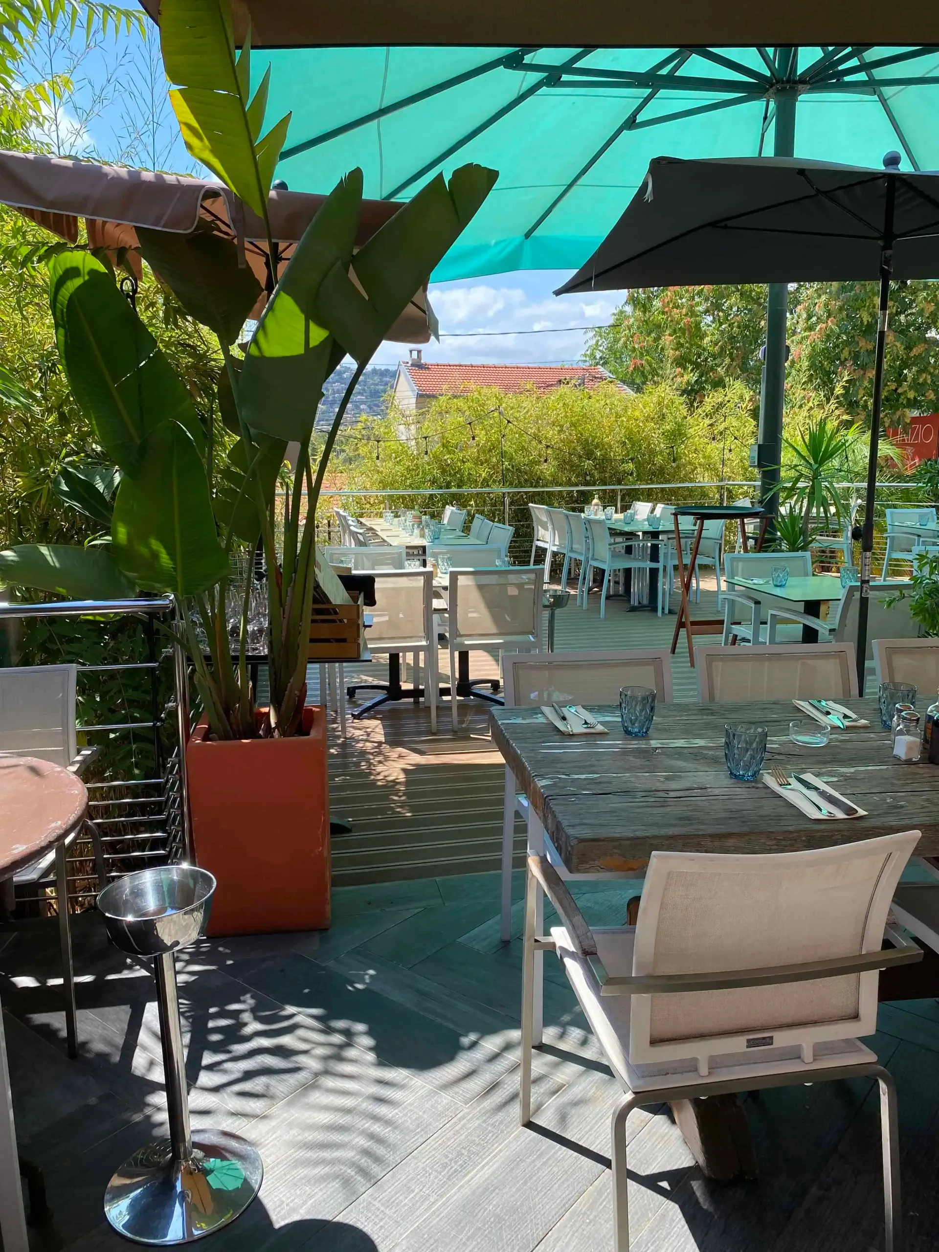 Terrasse extérieure de restaurant avec tables, chaises et plantes en pot sous des parasols verts et noirs par une journée ensoleillée.