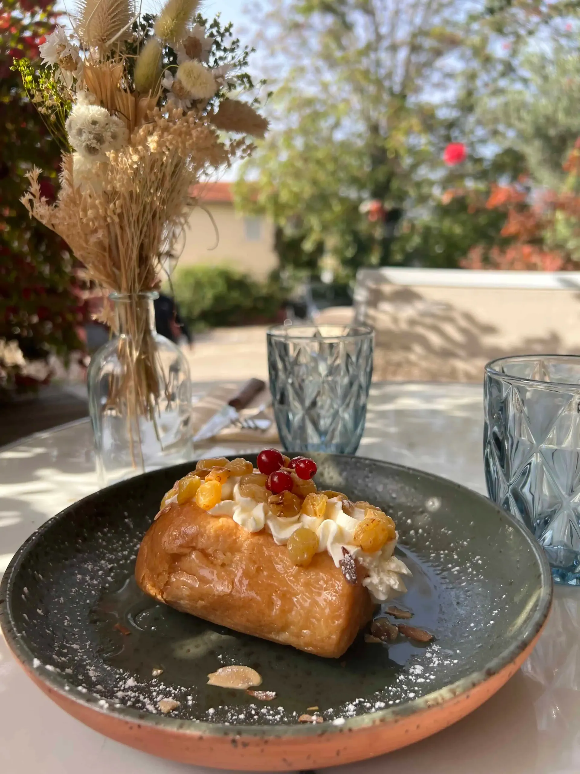Un gâteau baba garni de crème fouettée, de fruits jaunes et de groseilles rouges, servi sur une assiette grise sur une table en terrasse.