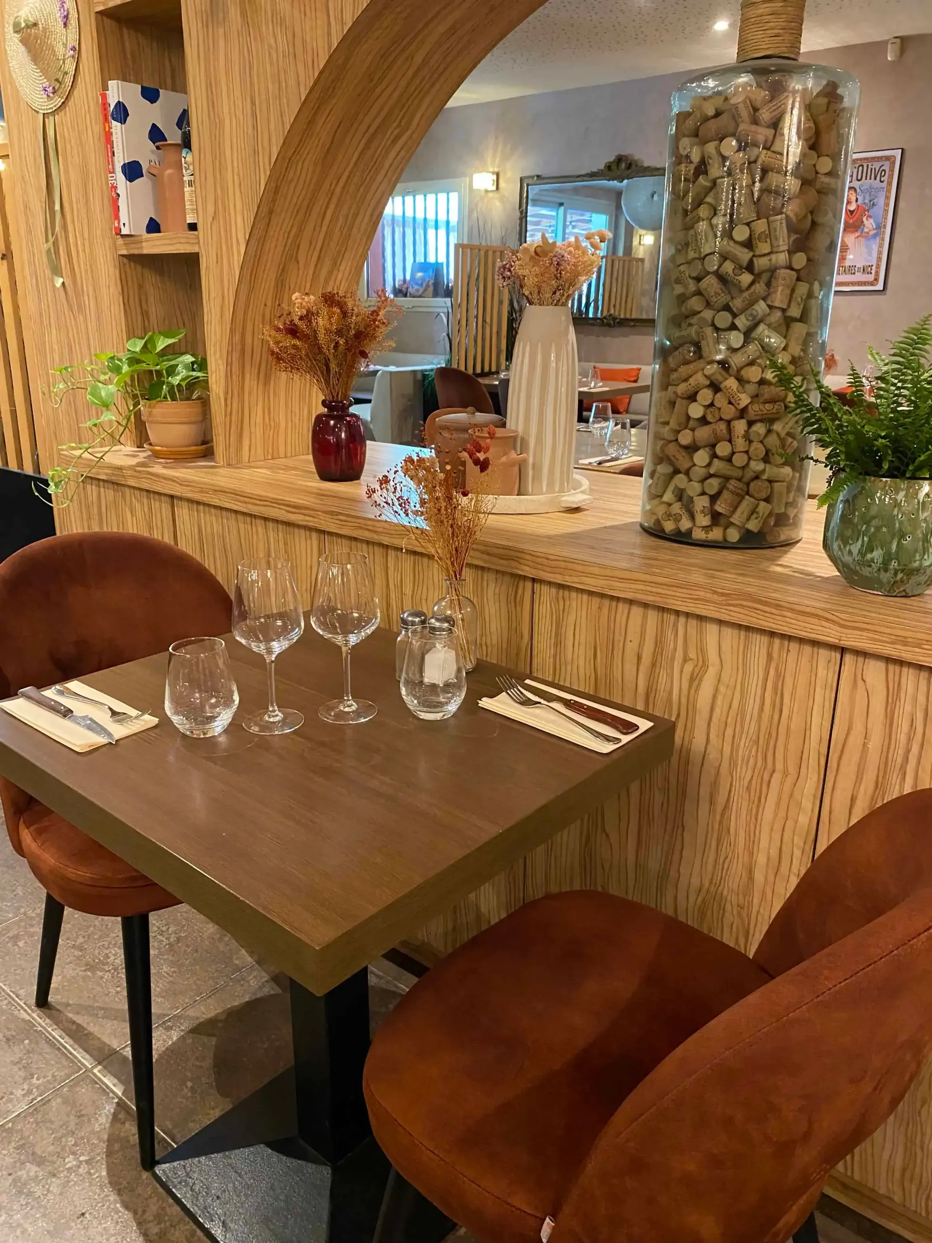 Une table de restaurant dressée pour deux, avec des chaises en velours marron, des verres à vin et des couverts, encadrée par une arche en bois.