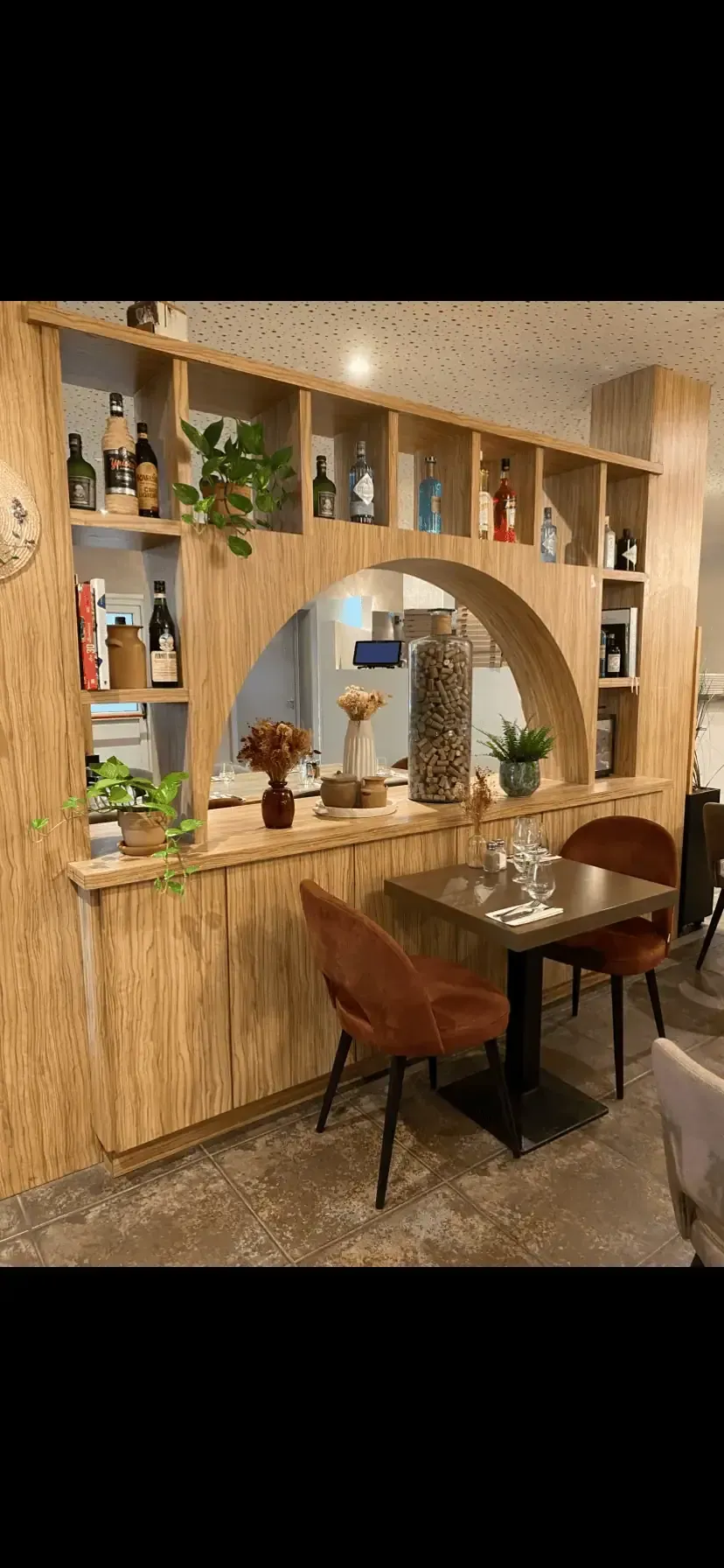 Une cloison cintrée en bois clair avec des étagères intégrées présentant des bouteilles et des plantes, à côté d'une petite table avec deux chaises.