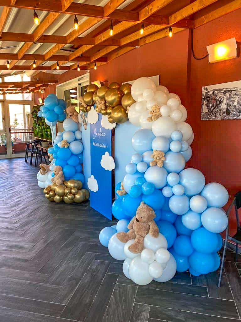 Arche de ballons décorative dans les tons bleus, blancs et dorés, ornée d'ours en peluche et d'un décor de fond, installée sur un sol en bois.